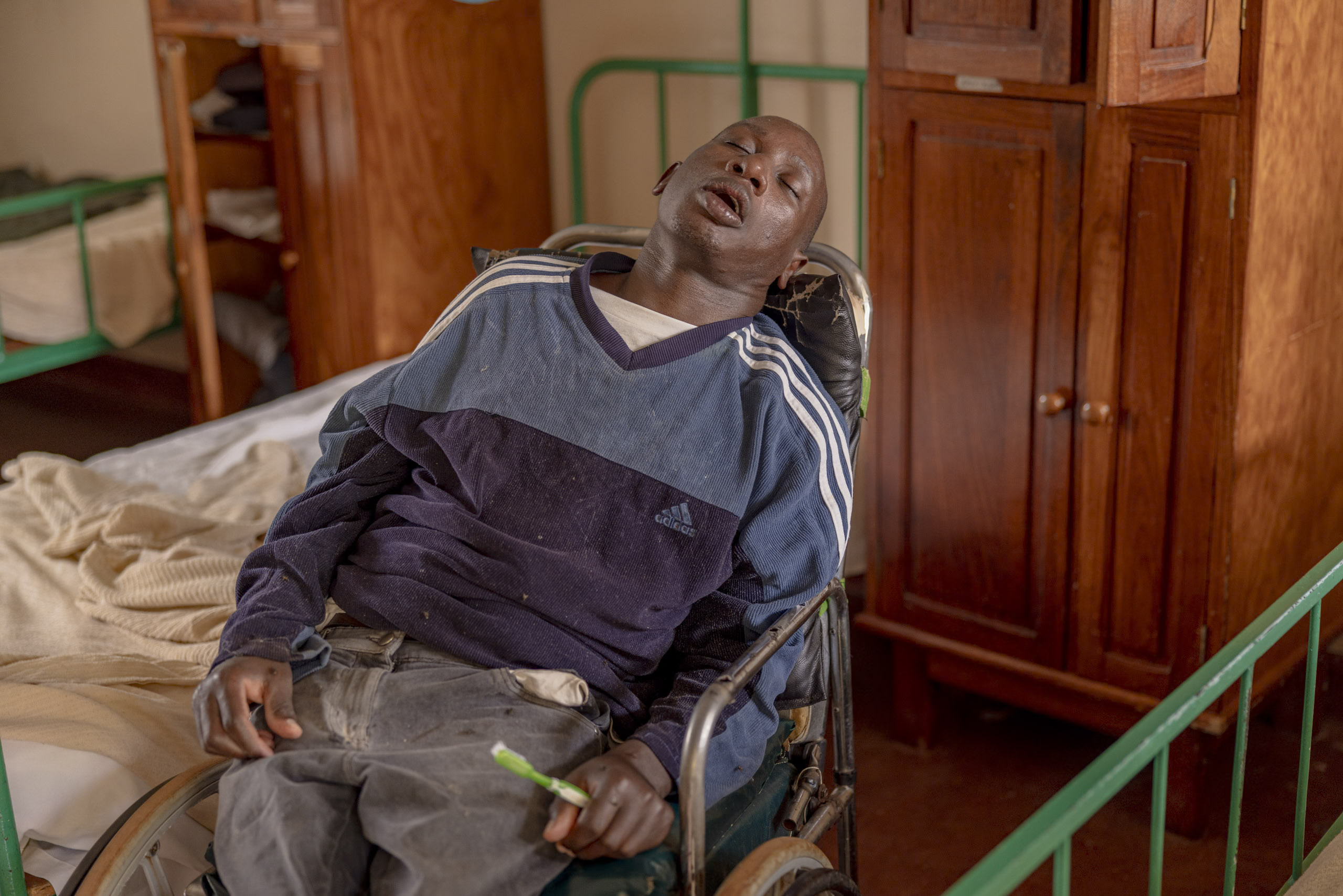 Ein Mann mit geschlossenen Augen, der einen gestreiften Pullover und Jeans trägt, lehnt sich in einem Rollstuhl in einem Zimmer zurück und hält eine Zahnbürste in der Hand. © Fotografie Tomas Rodriguez