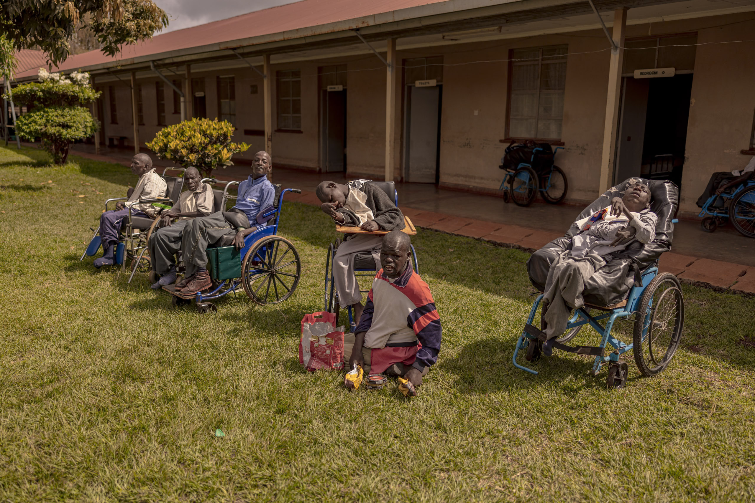 Eine Gruppe Erwachsener in Rollstühlen versammelte sich vor einem Gebäude mit Rasen und widmete sich unter einem strahlend sonnigen Himmel verschiedenen Aktivitäten. © Fotografie Tomas Rodriguez