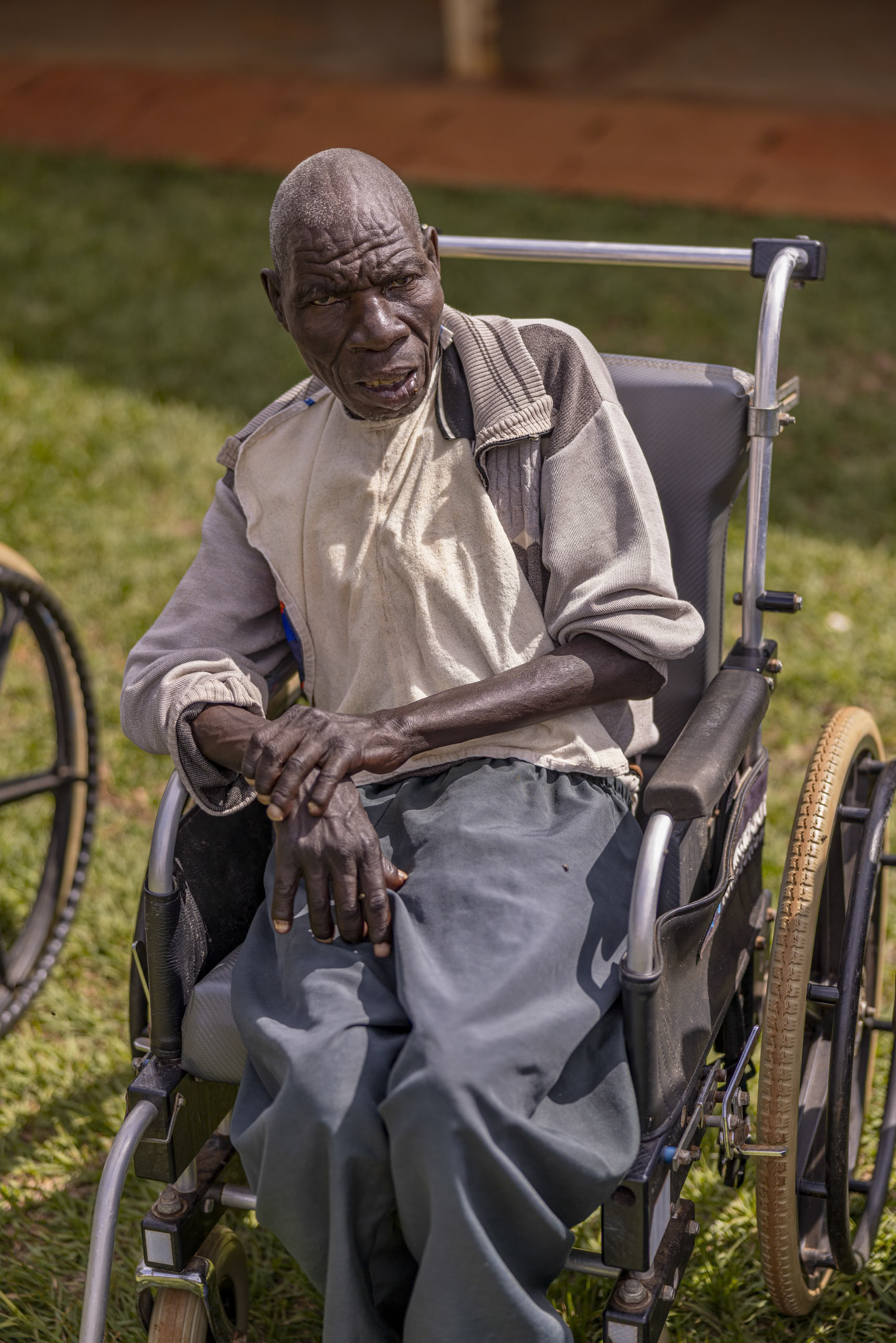 Ein älterer Mann sitzt nachdenklich in einem Rollstuhl im Freien, bekleidet mit einem beigen Hemd und einer grauen Hose, umgeben von einer grünen Rasenfläche. © Fotografie Tomas Rodriguez