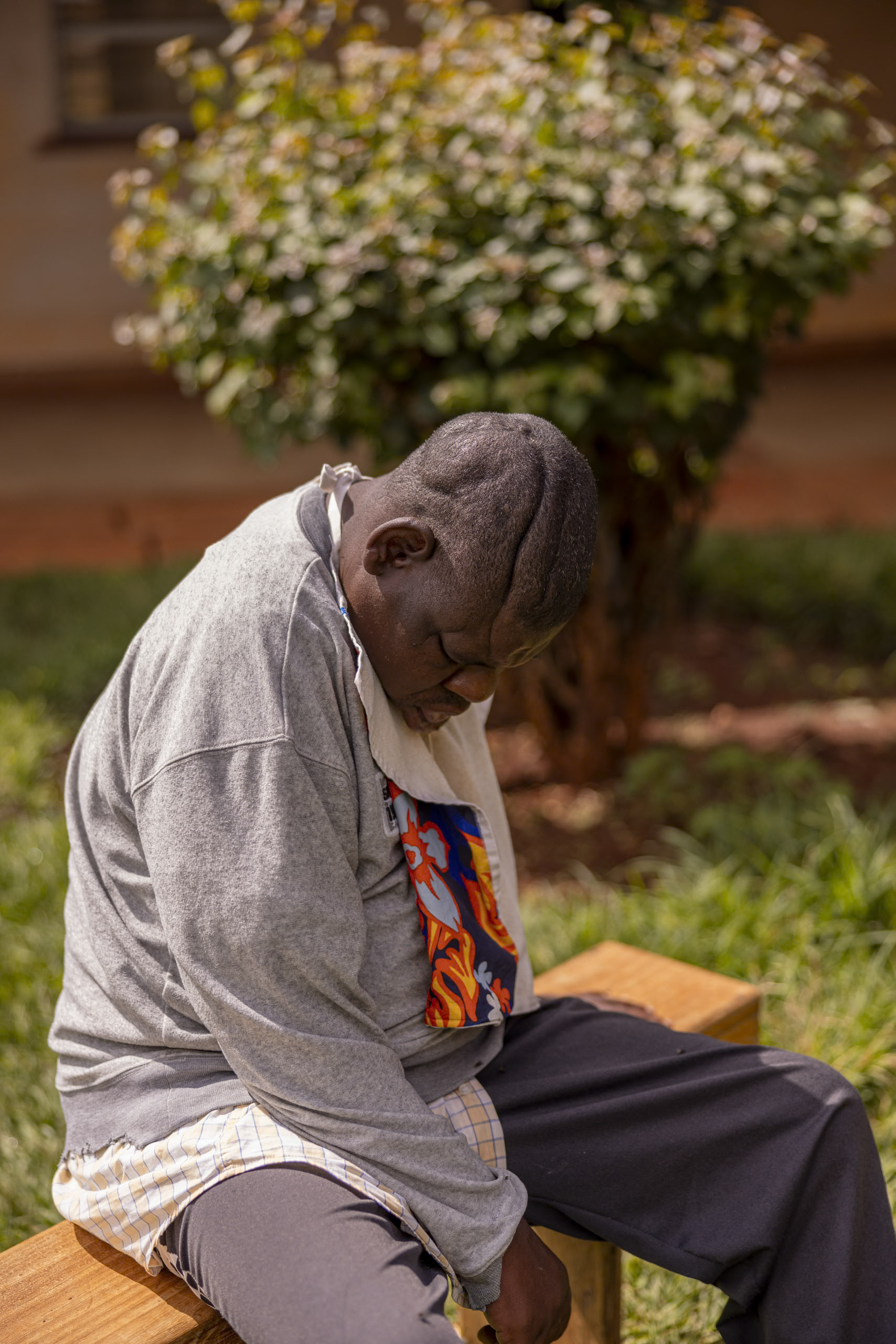 Ein älterer Mann sitzt ruhig auf einer Holzbank, den Kopf nachdenklich gesenkt, im Hintergrund ein blühender Strauch und üppiges Gras. Er trägt ein graues Sweatshirt und ein gemustertes Hemd. © Fotografie Tomas Rodriguez