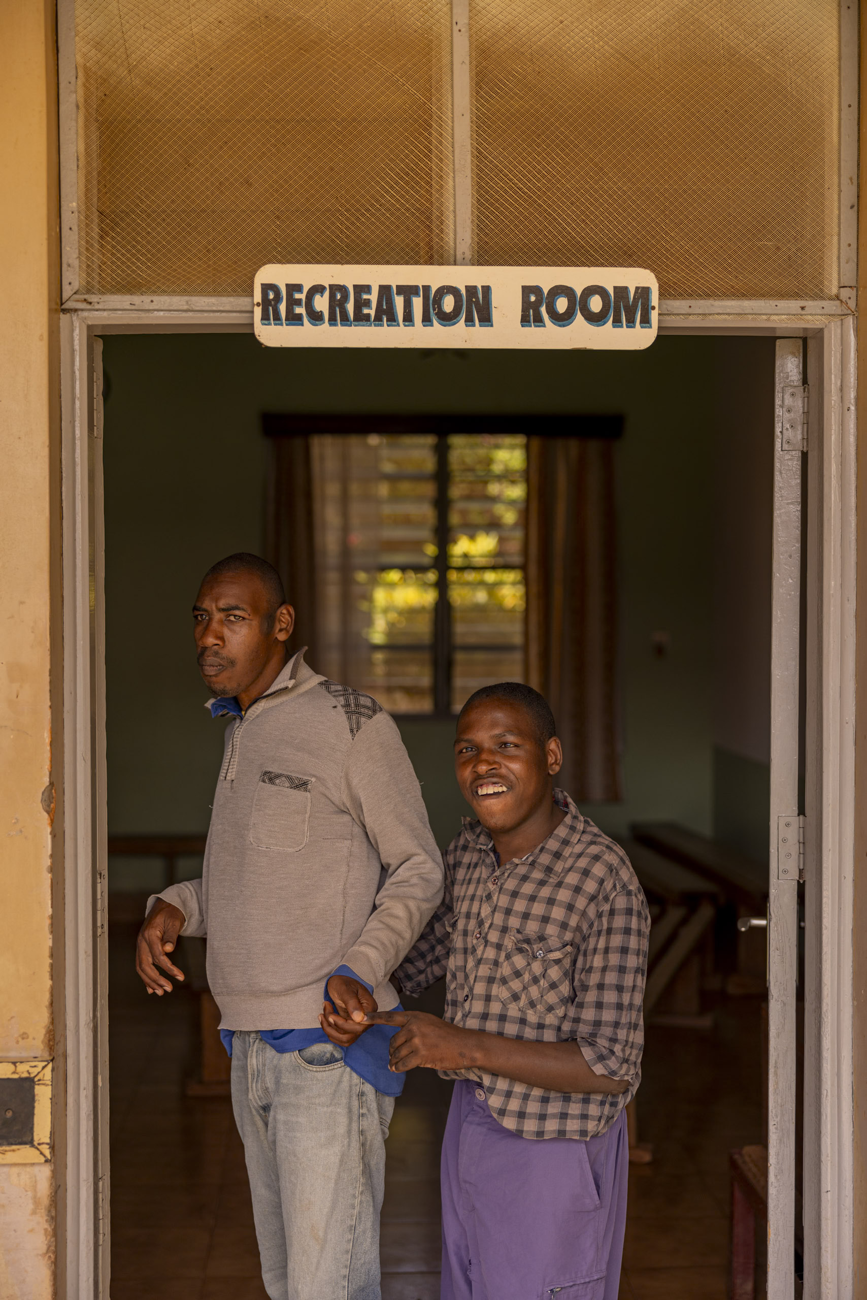 Zwei Männer, einer stehend, der andere im Rollstuhl, lächeln im Türrahmen eines „Aufenthaltsraums“ in einem warm beleuchteten Gebäude in die Kamera. © Fotografie Tomas Rodriguez