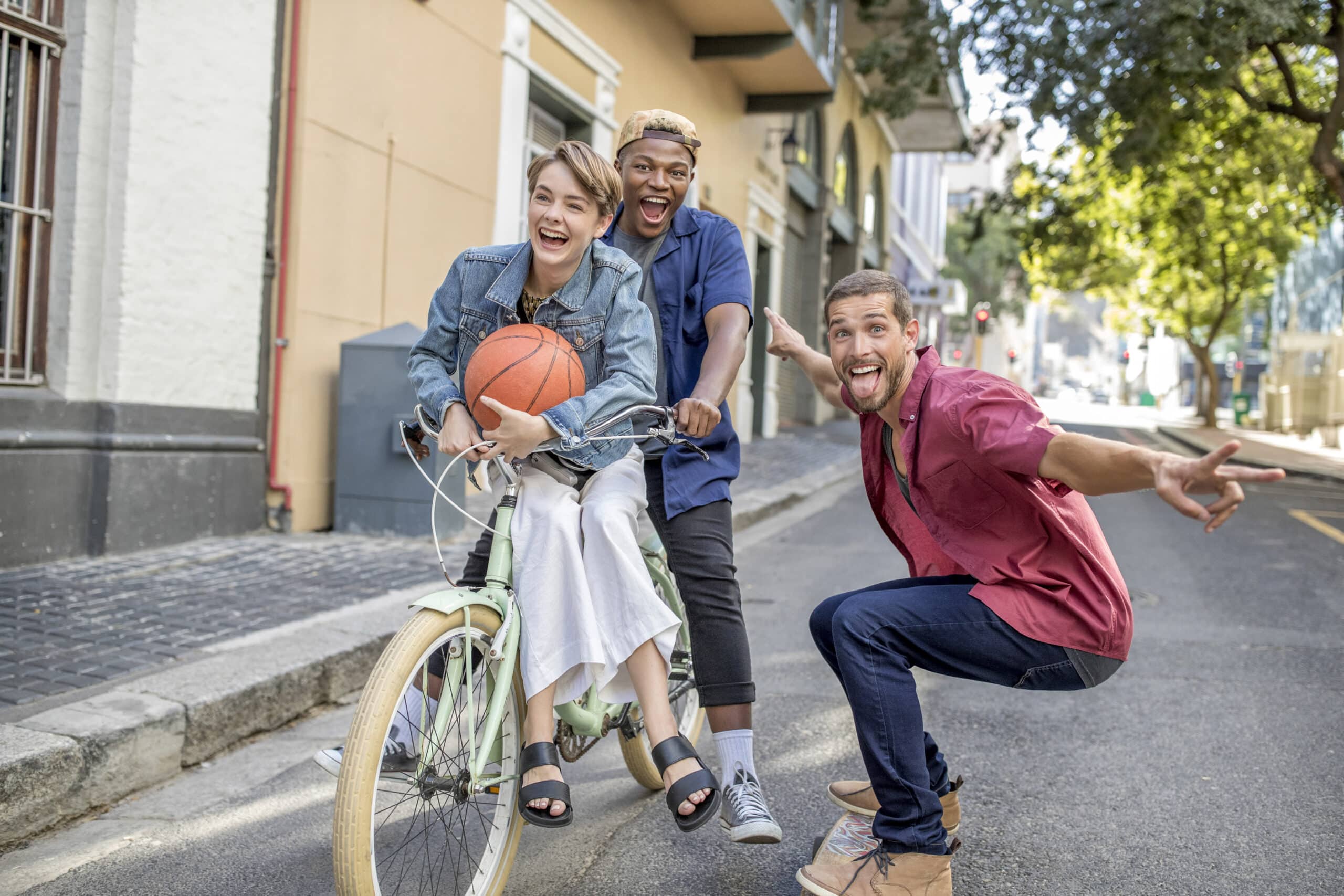 Drei Freunde genießen einen lustigen Moment im Freien. Eine Person sitzt mit einem Basketball auf einem Fahrrad, während eine andere fährt, und eine dritte macht eine verspielte Pose neben ihnen. Die Straße und die Bäume bilden eine lebendige Kulisse für ihre Mätzchen. © Fotografie Tomas Rodriguez
