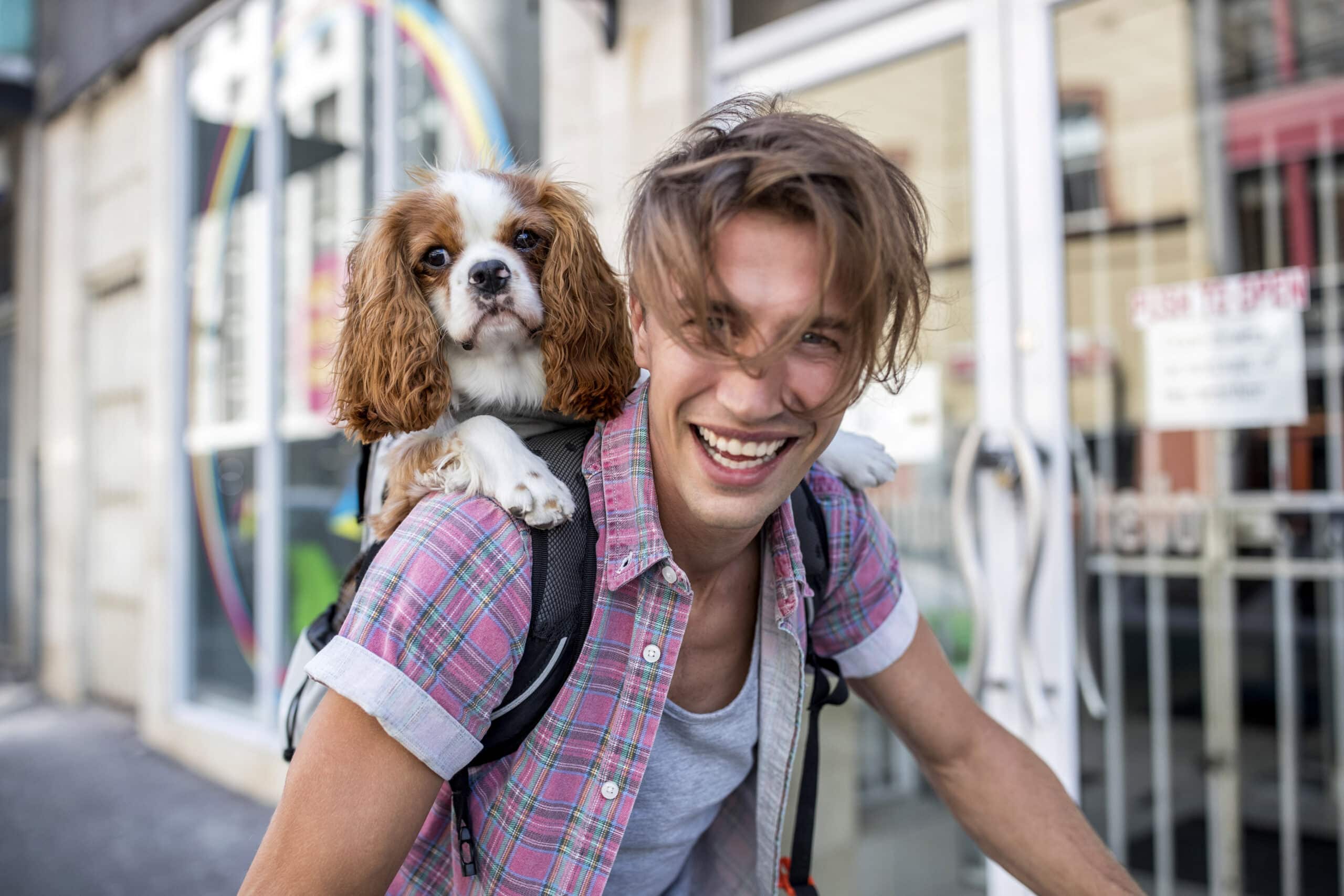 Ein lächelnder Mann mit zerzaustem Haar, einem karierten Hemd und einem Rucksack radelt auf einer Stadtstraße. Ein kleiner Hund mit langen Ohren lugt aus dem Rucksack hervor und genießt die Fahrt. Der Hintergrund ist verschwommen, zeigt aber Gebäude und ein farbenfrohes Wandgemälde. © Fotografie Tomas Rodriguez