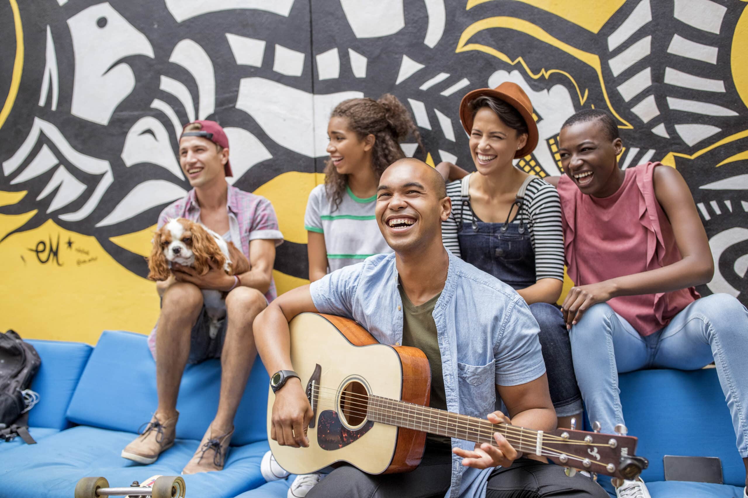 Eine Gruppe von fünf Freunden sitzt auf einem blauen Sofa vor einem farbenfrohen Wandgemälde. Eine Person in der ersten Reihe klimpert auf einer Gitarre, während eine andere einen Hund hält. Sie alle lächeln und lachen und genießen den Moment zusammen. © Fotografie Tomas Rodriguez
