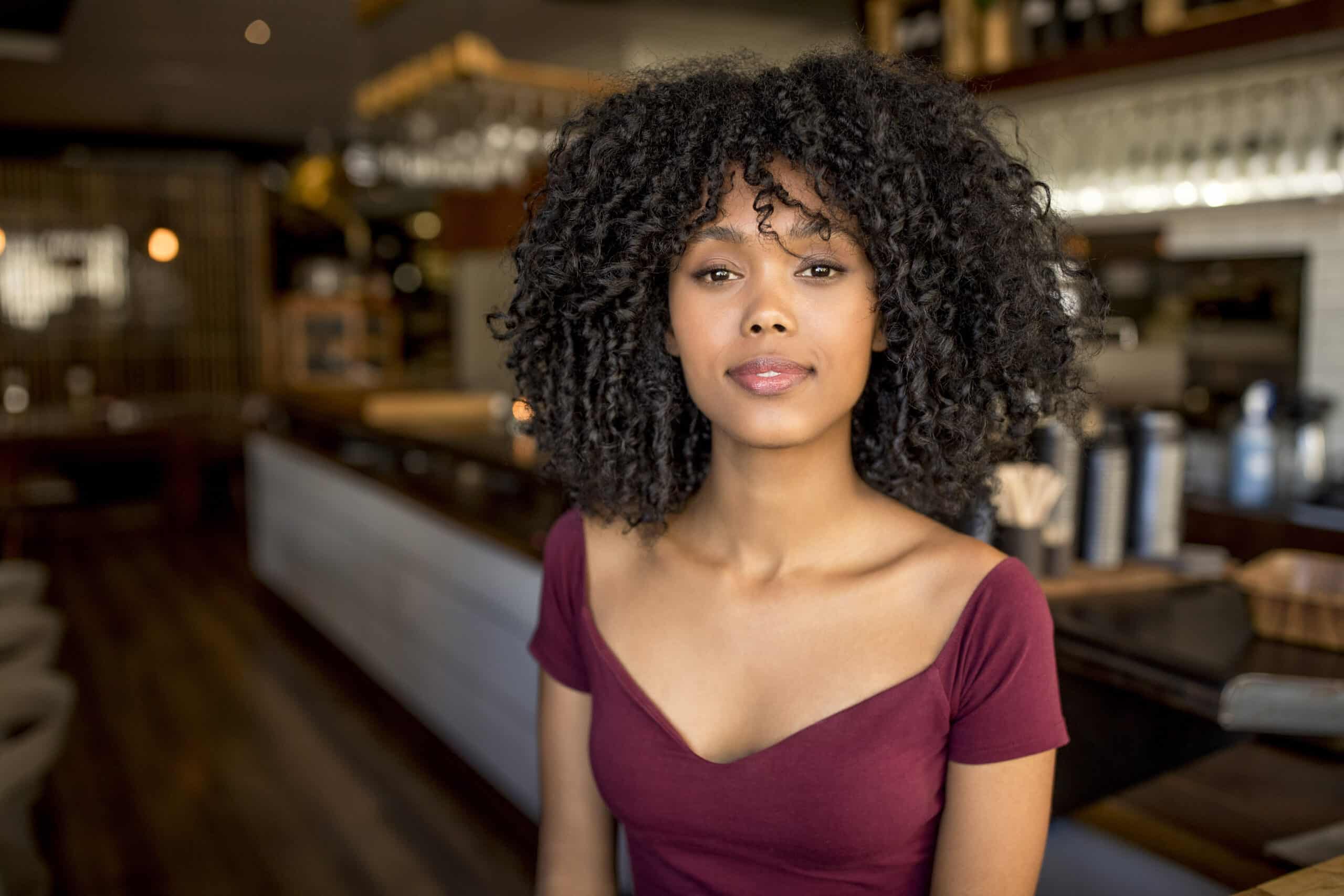 Eine Frau mit lockigem Haar und einem schulterfreien, kastanienbraunen Top steht in einem schwach beleuchteten Café oder Restaurant. Sie blickt direkt in die Kamera, im Hintergrund sind Regale und hängende Gläser zu sehen. Das Ambiente wirkt gemütlich und einladend. © Fotografie Tomas Rodriguez
