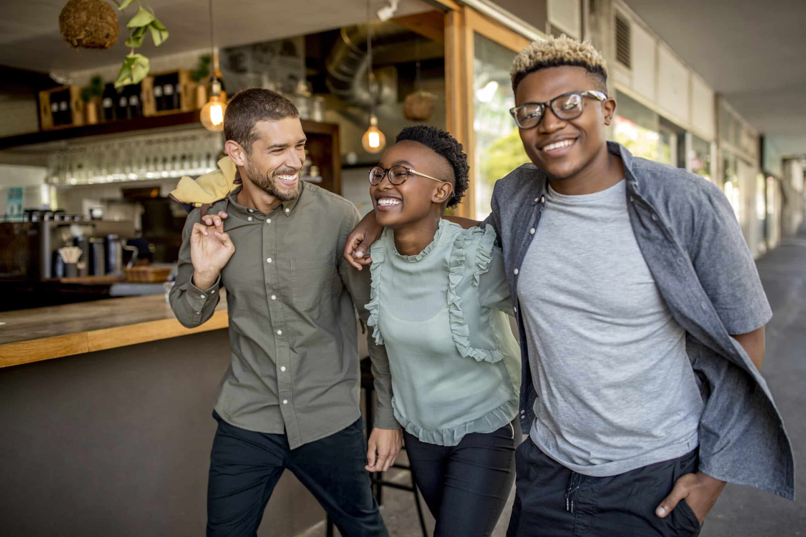Drei Freunde gehen zusammen spazieren und lächeln, zwei Männer und eine Frau, die sich umarmen. Sie sind leger gekleidet und scheinen die Gesellschaft des anderen in der Nähe eines Cafés oder einer Markthalle zu genießen. Warmes und legeres Ambiente mit Hängelampen im Hintergrund. © Fotografie Tomas Rodriguez