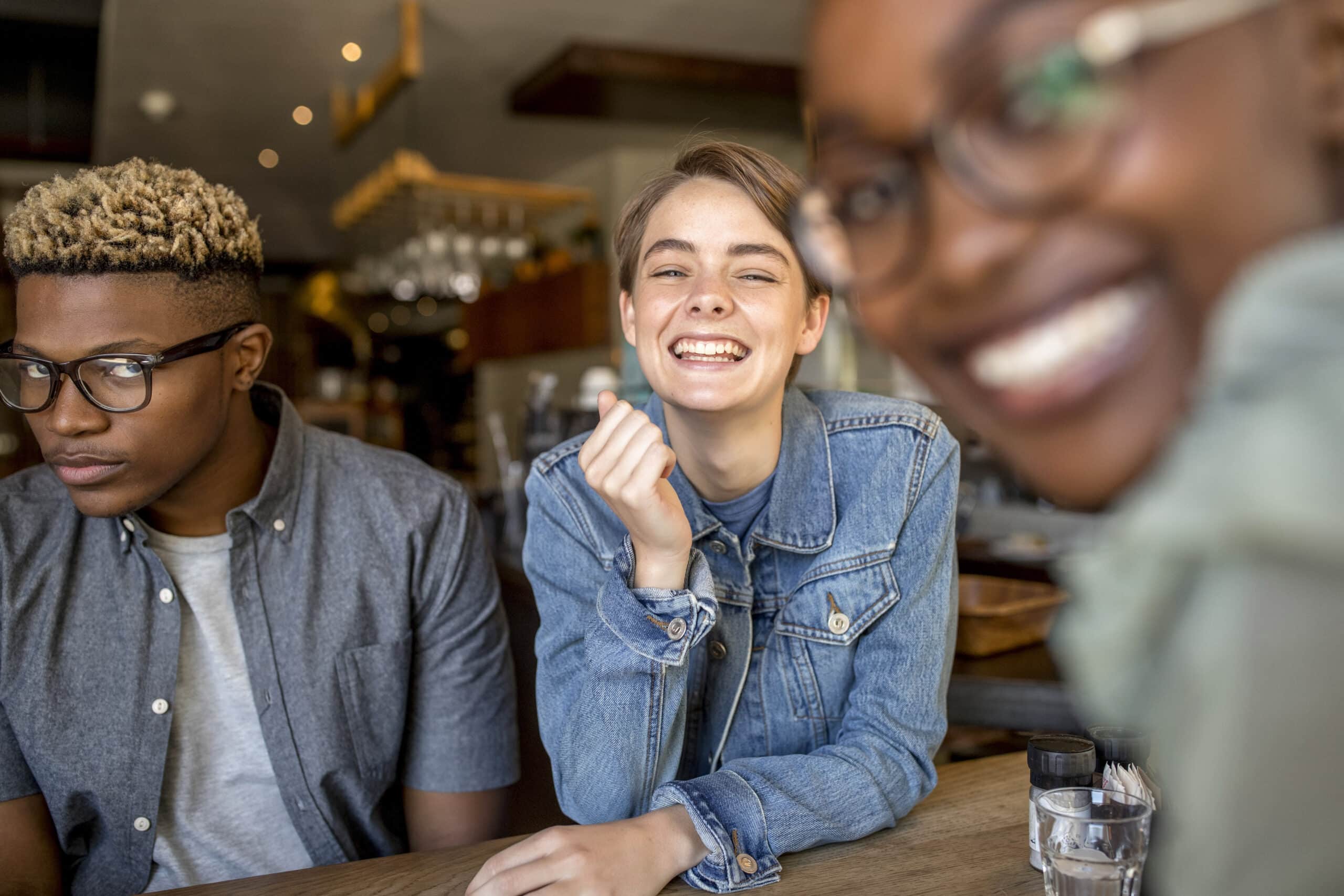 Drei Freunde sitzen an einem Cafétisch. Eine Person mit kurzen hellen Haaren und einer Jeansjacke lächelt strahlend in die Kamera. Eine andere Person mit kurzen blonden Haaren und Brille schaut mit neutralem Gesichtsausdruck nach unten. Eine dritte Person mit Brille ist unscharf im Vordergrund zu sehen. © Fotografie Tomas Rodriguez