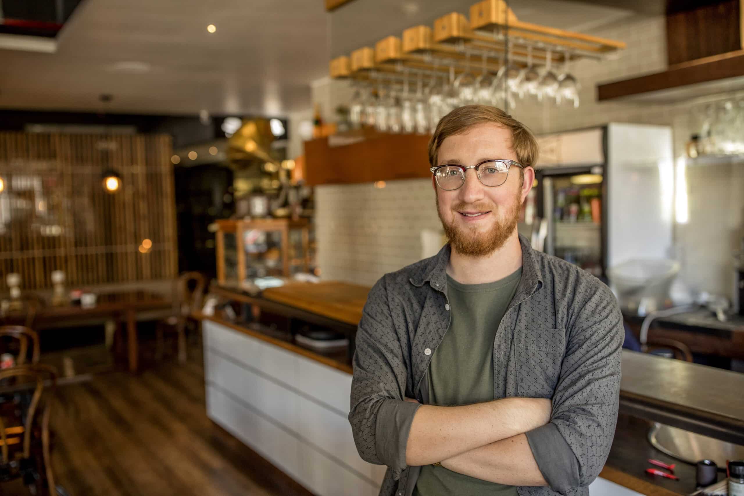 Ein Mann mit Brille und Bart steht lächelnd mit verschränkten Armen in einem gemütlichen, modernen Café. Das Café verfügt über eine Holztheke, hängende Weingläser und eine warme, einladende Atmosphäre mit Holzmöbeln und sanfter Beleuchtung. © Fotografie Tomas Rodriguez