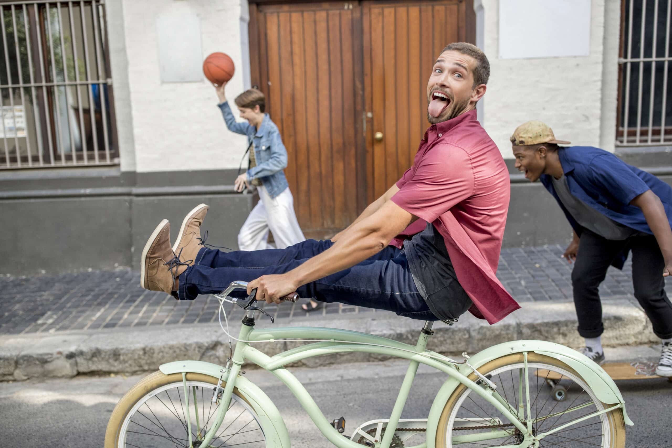 Im Fokus steht ein Mann auf einem grünen Fahrrad, dessen Beine nach vorne gestreckt sind und dessen Zunge heraushängt. Hinter ihm greift ein anderer Mann nach einem Basketball in der Luft, während sich ein dritter Mann bückt, wahrscheinlich um zu spielen oder etwas von der Straße aufzuheben. © Fotografie Tomas Rodriguez