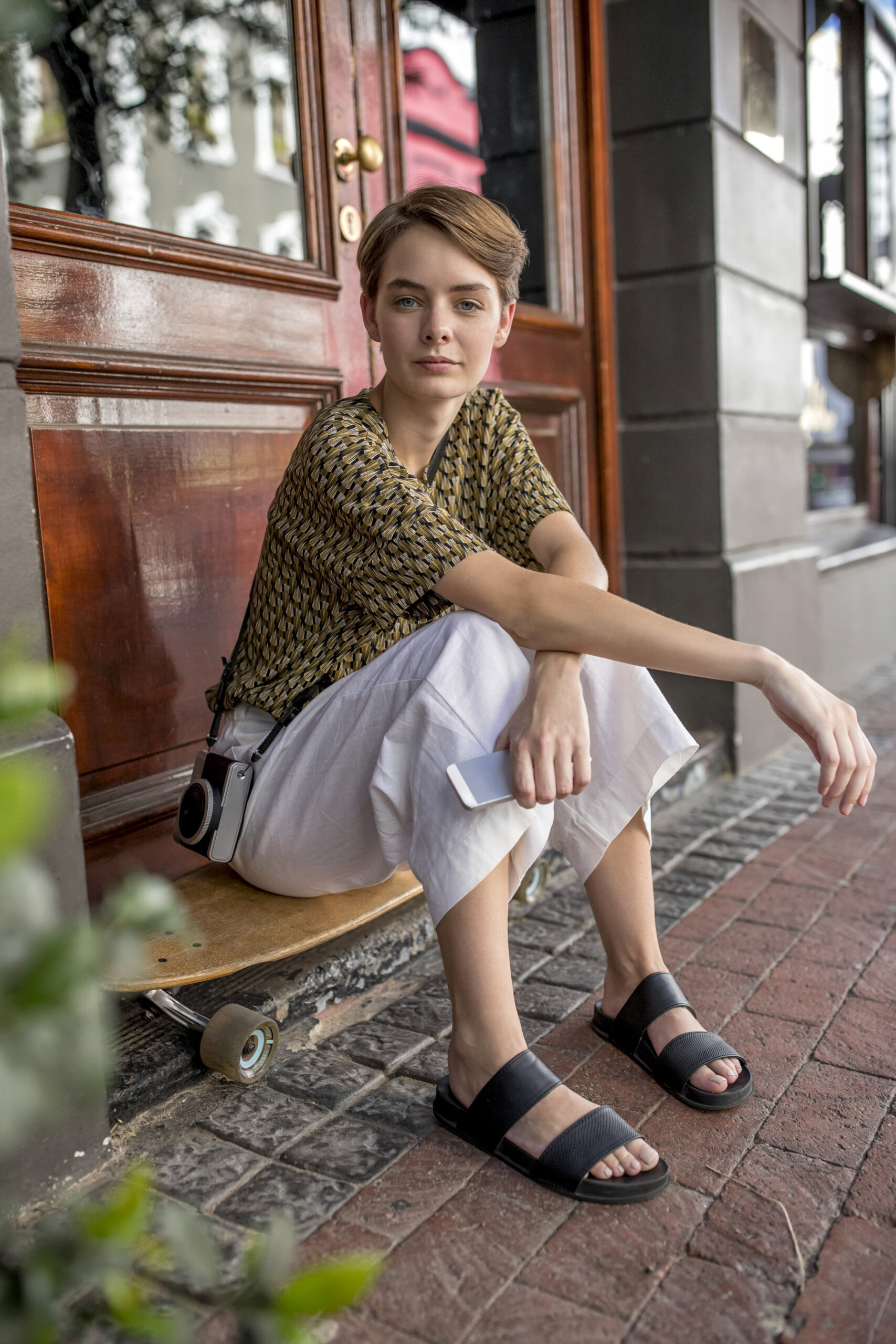 Eine Person mit kurzen Haaren sitzt auf einem Skateboard vor einem Gebäude mit Holztüren und hält ein Smartphone in der Hand. Sie trägt ein gemustertes Hemd, weiße Hosen, schwarze Sandalen und eine Kamera um die Schulter. Die Szenerie scheint eine Stadtstraße zu sein. © Fotografie Tomas Rodriguez