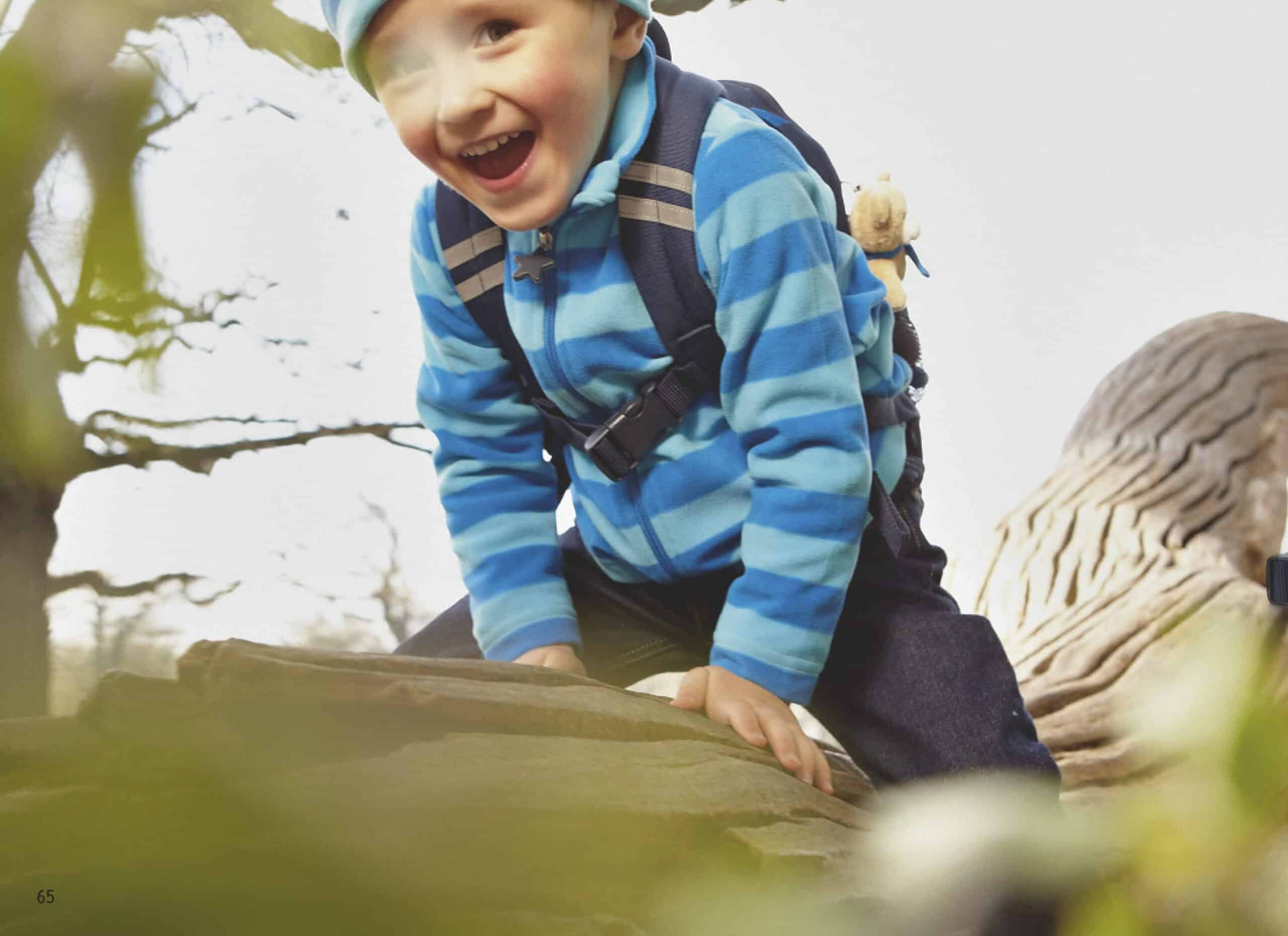 Ein fröhlicher kleiner Junge in einer blau gestreiften Jacke und einem Rucksack klettert im Freien auf einen großen Felsen und lächelt breit. © Fotografie Tomas Rodriguez