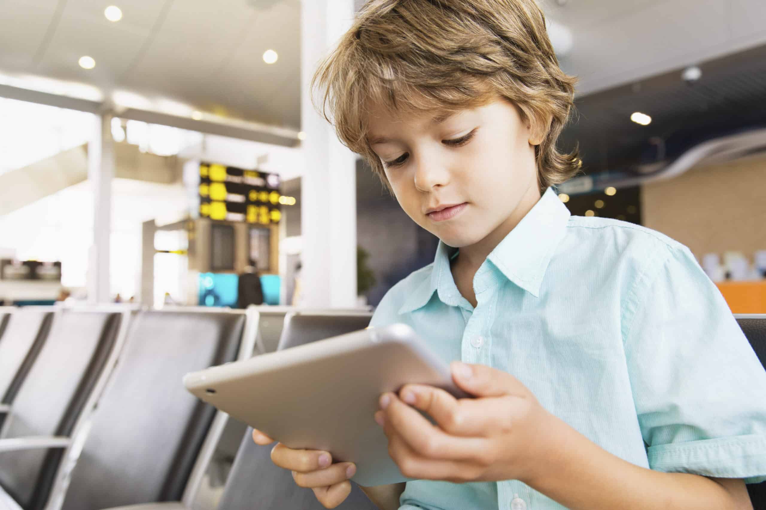Ein kleines Kind mit blonden Haaren und einem hellblauen Hemd sitzt in einem Flughafenterminal und hält ein Tablet in der Hand. Das Kind scheint auf den Bildschirm zu starren. Im Hintergrund sind Stuhlreihen und eine Anzeigetafel mit Fluginformationen zu sehen. © Fotografie Tomas Rodriguez