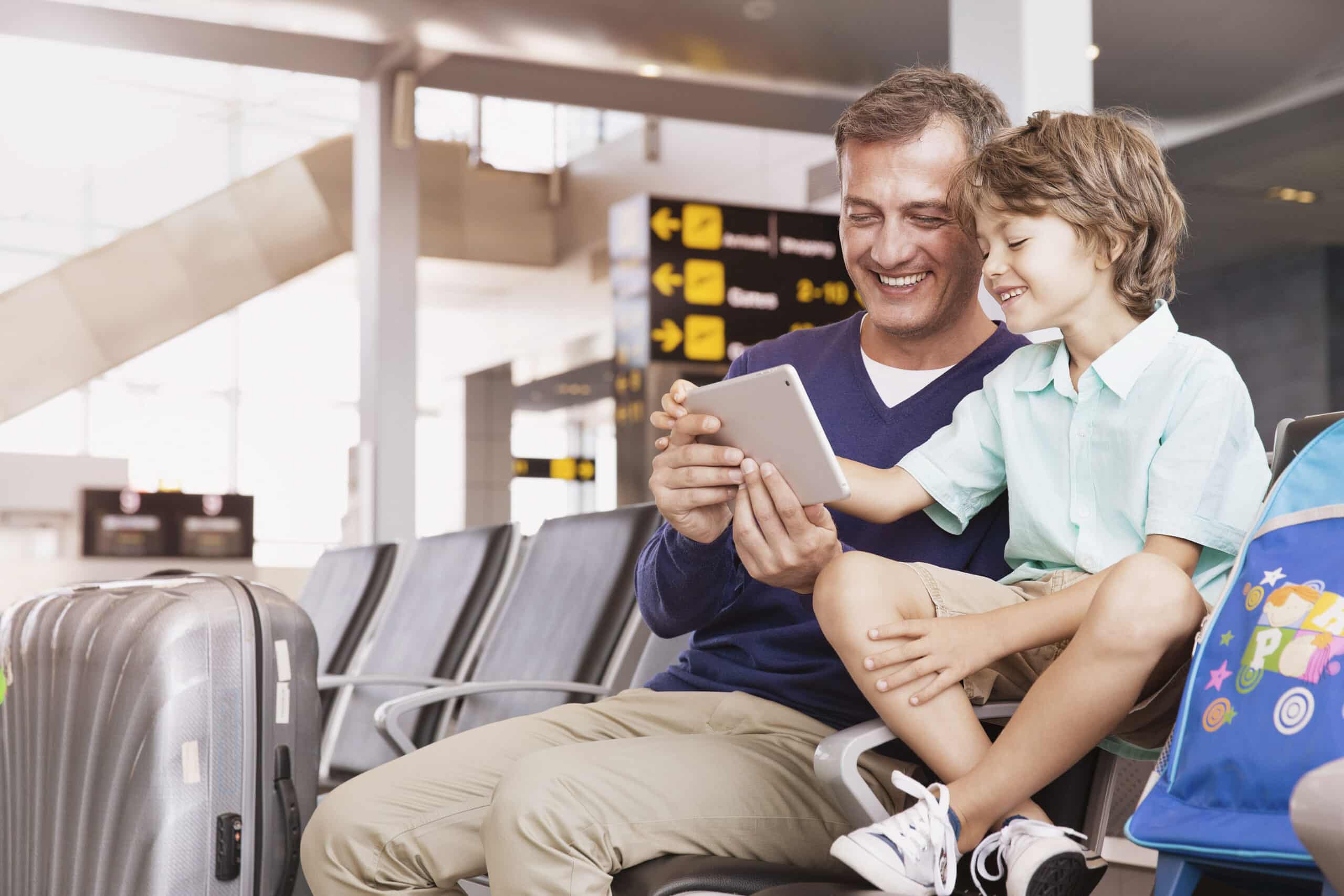 Ein Mann und ein kleiner Junge sitzen in einem Flughafenterminal, lächeln und schauen gemeinsam auf ein Tablet. Der Mann trägt einen blauen Pullover und der Junge ein hellgrünes Hemd. In der Nähe stehen Koffer und ein blauer Rucksack. Im Hintergrund sind Schilder mit Richtungspfeilen zu sehen. © Fotografie Tomas Rodriguez