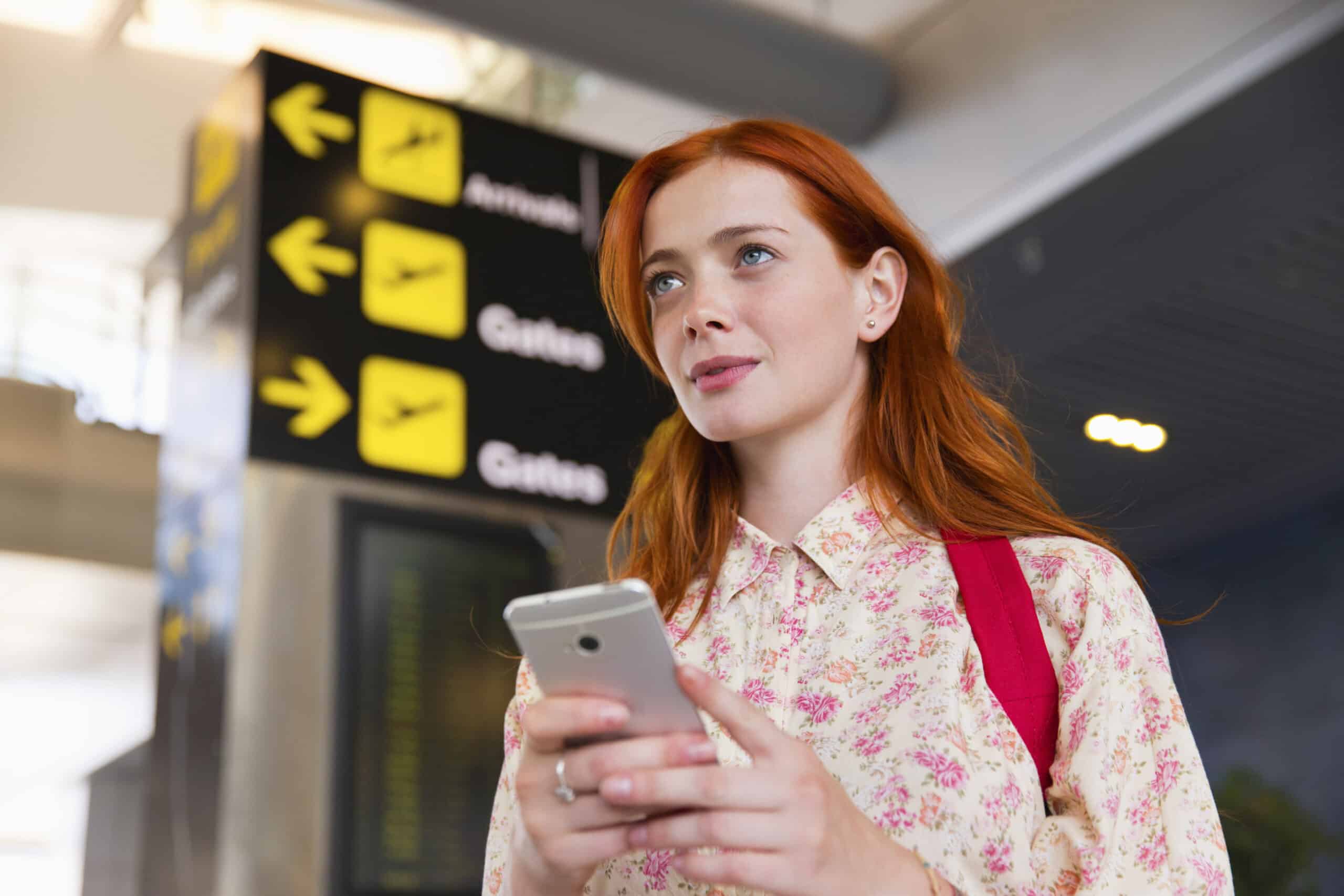 Eine Frau mit langen roten Haaren, die ein Hemd mit Blumenmuster und einen roten Rucksack trägt, hält ein Smartphone in der Hand. Hinter ihr ist ein Flughafenschild mit Pfeilen zu Ankünften, Gates und anderen Zielen zu sehen. Sie scheint zu warten oder nach dem Weg zu suchen. © Fotografie Tomas Rodriguez