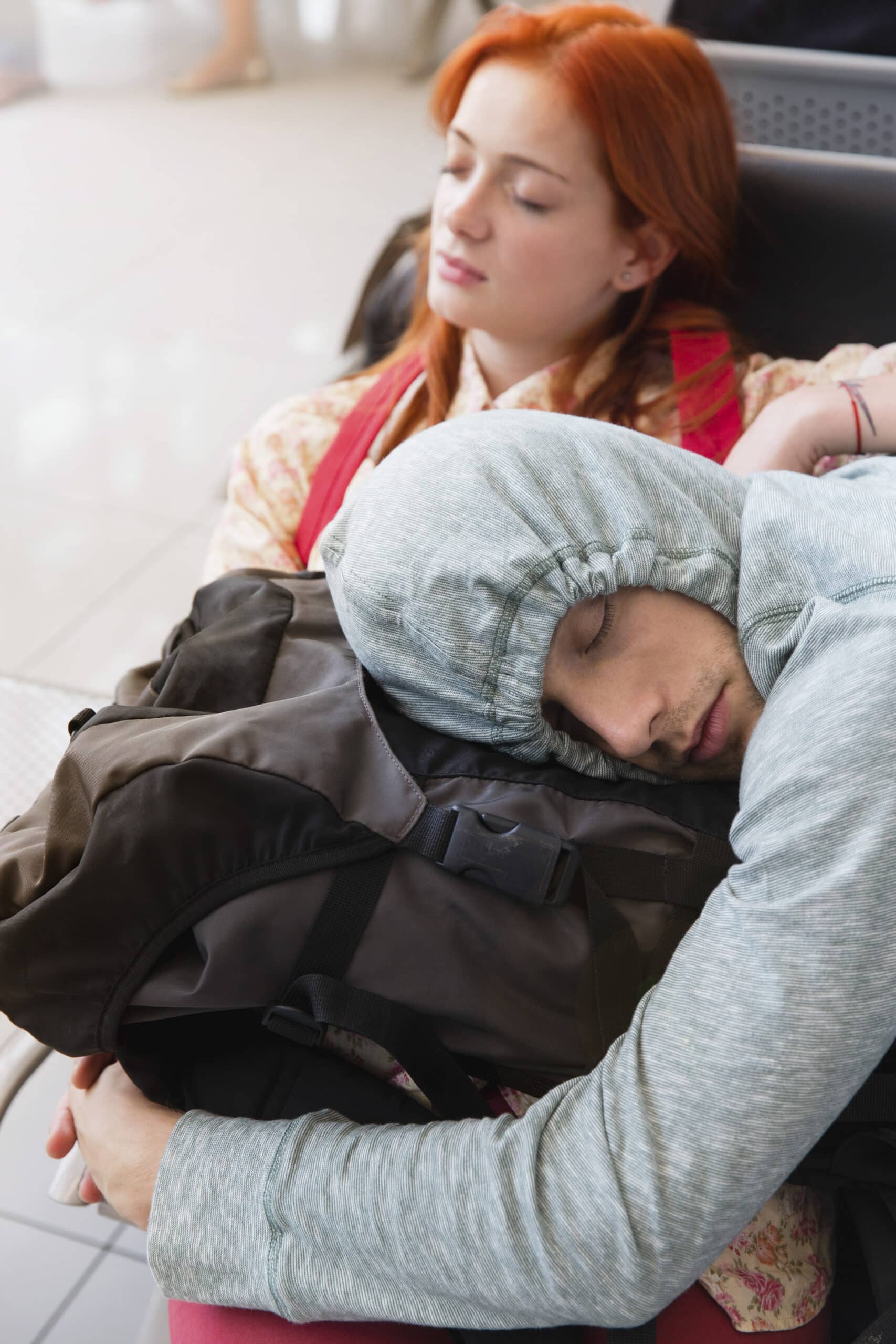 Zwei junge Reisende schlafen auf einem Flughafen. Der Mann im Vordergrund trägt einen grauen Kapuzenpullover mit hochgezogener Kapuze und ruht mit seinem Kopf auf einem schwarzen Rucksack. Hinter ihm schläft eine Frau mit roten Haaren und einem hellen Jäckchen, das ihren Kopf in den Nacken legt. © Fotografie Tomas Rodriguez