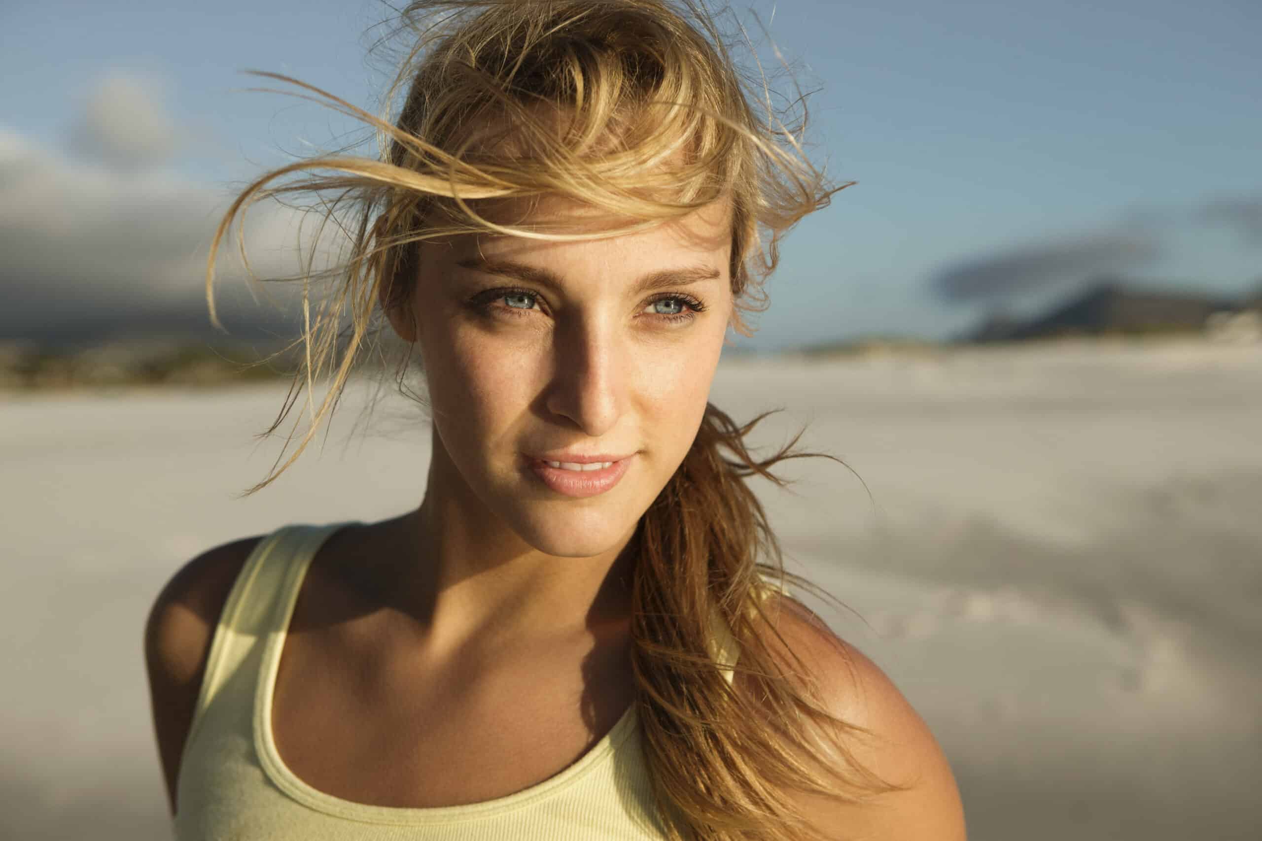 Eine junge Frau mit im Wind wehendem blonden Haar und einem gelben Tanktop steht an einem Sandstrand vor einem unscharfen Hintergrund. © Fotografie Tomas Rodriguez