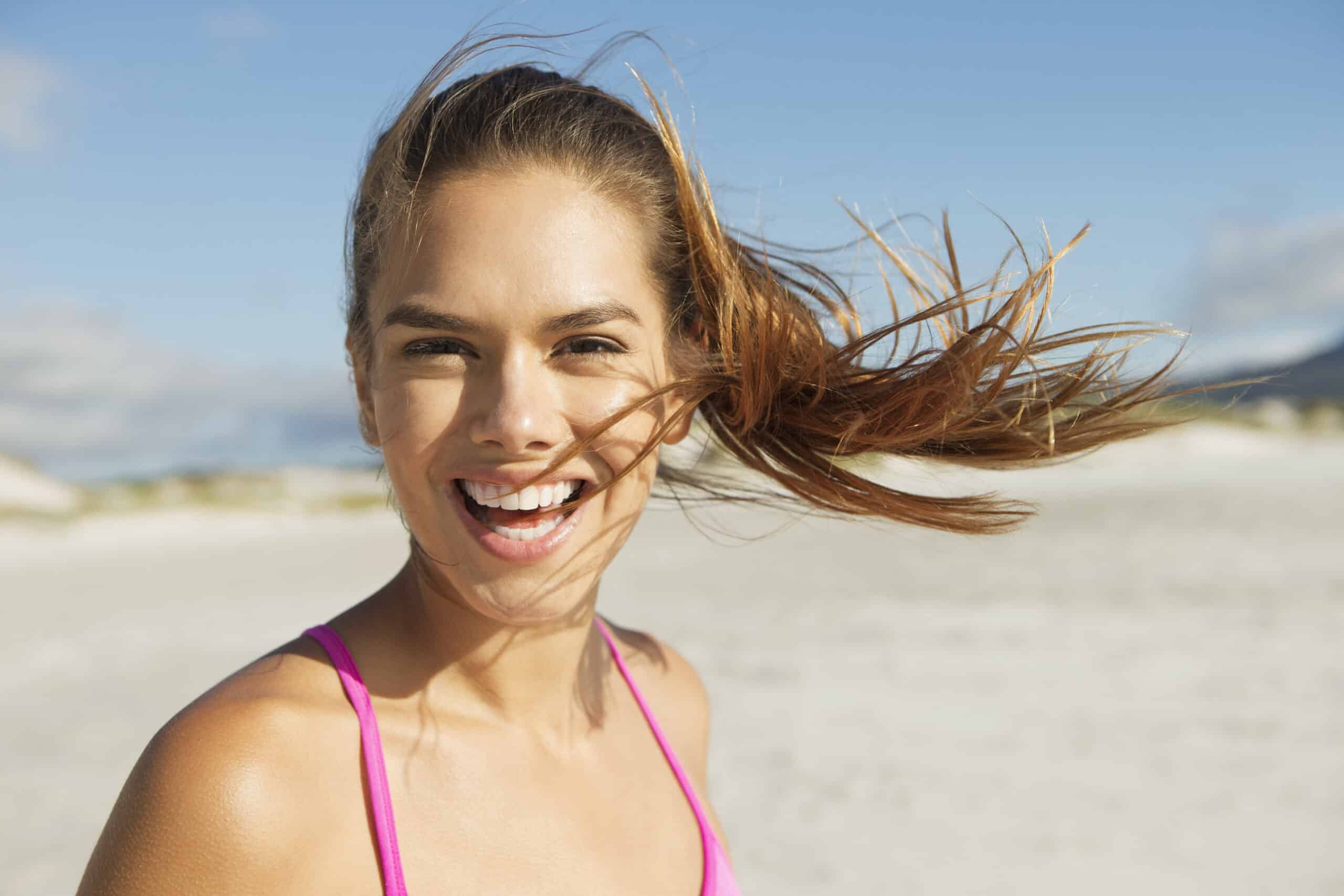 Eine fröhliche junge Frau mit wallendem braunem Haar lächelt breit an einem sonnigen Strand und trägt ein rosa Tanktop. Ihr Haar weht in der Brise vor dem Hintergrund eines klaren Himmels und eines sandigen Ufers. © Fotografie Tomas Rodriguez