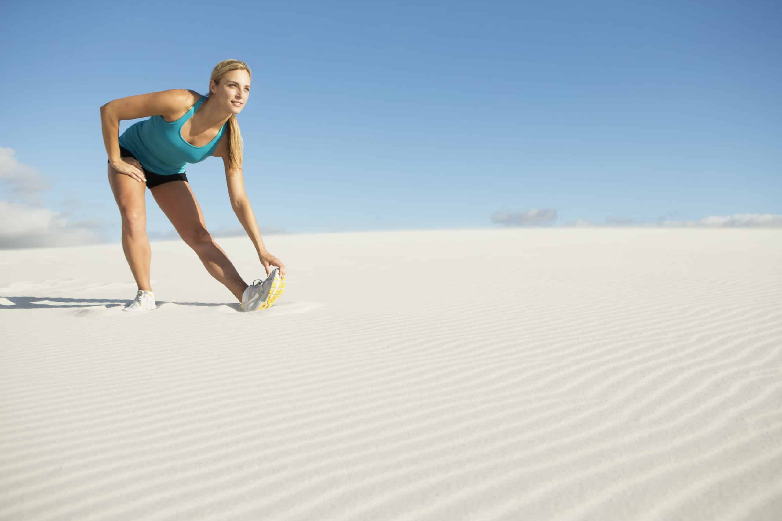 Eine Frau in Sportkleidung streckt sich unter einem strahlenden Himmel an einem Sandstrand, berührt mit einer Hand ihre Zehen und blickt nach vorne. © Fotografie Tomas Rodriguez