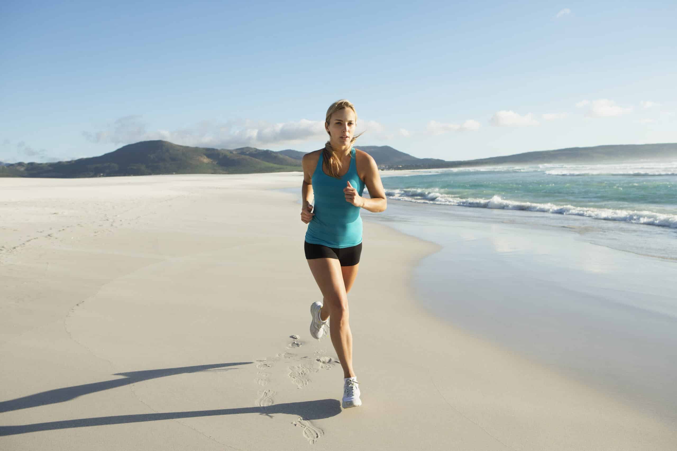 Eine Frau joggt an einem sonnigen Strand mit Bergen im Hintergrund. Sie trägt ein blaues Tanktop und schwarze Shorts und blickt nach vorne gerichtet, während sie am Ufer entlang läuft. © Fotografie Tomas Rodriguez