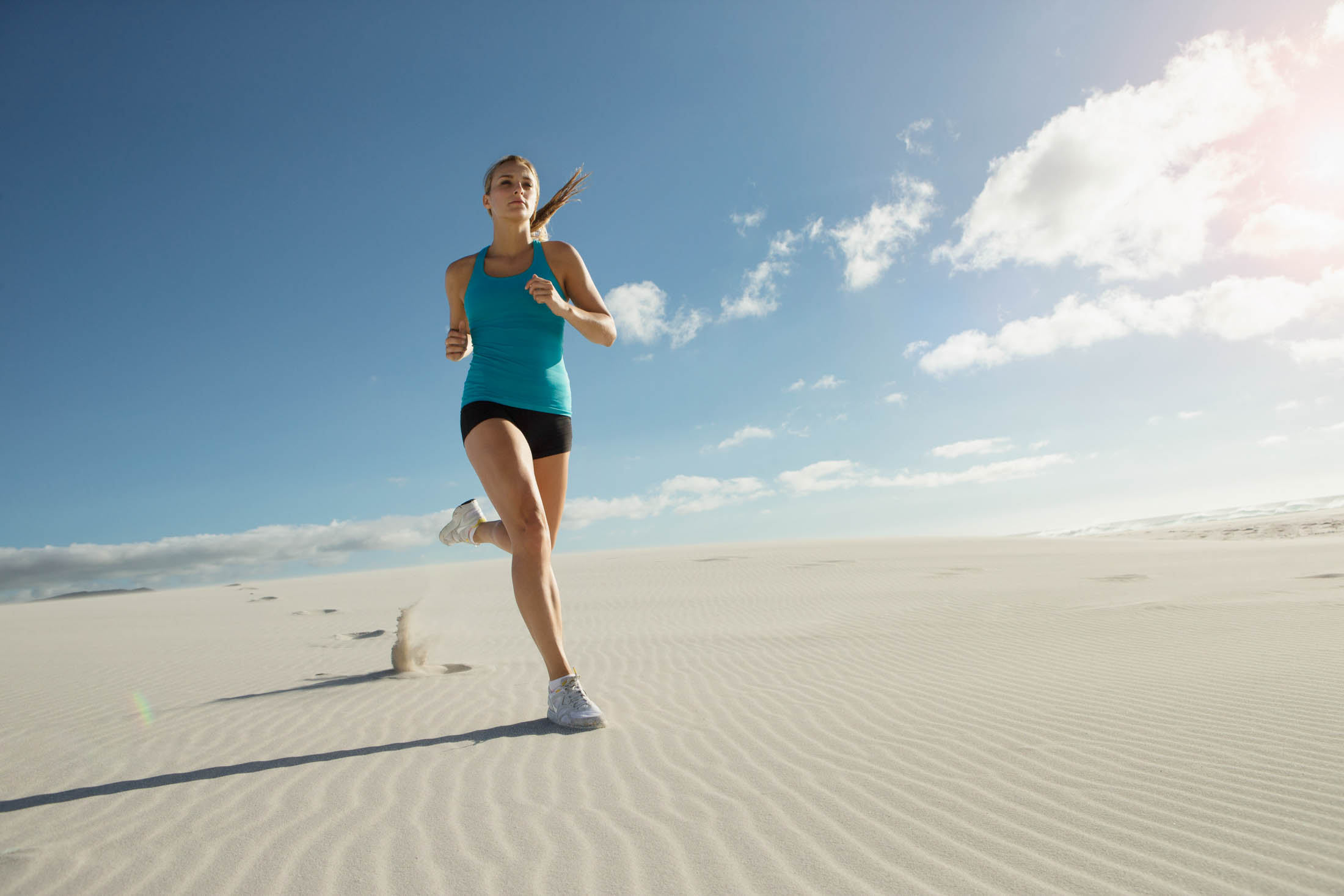 Eine Frau läuft unter klarem Himmel durch eine Sandwüste. Sie trägt ein blaues Tanktop, schwarze Shorts und weiße Laufschuhe. © Fotografie Tomas Rodriguez