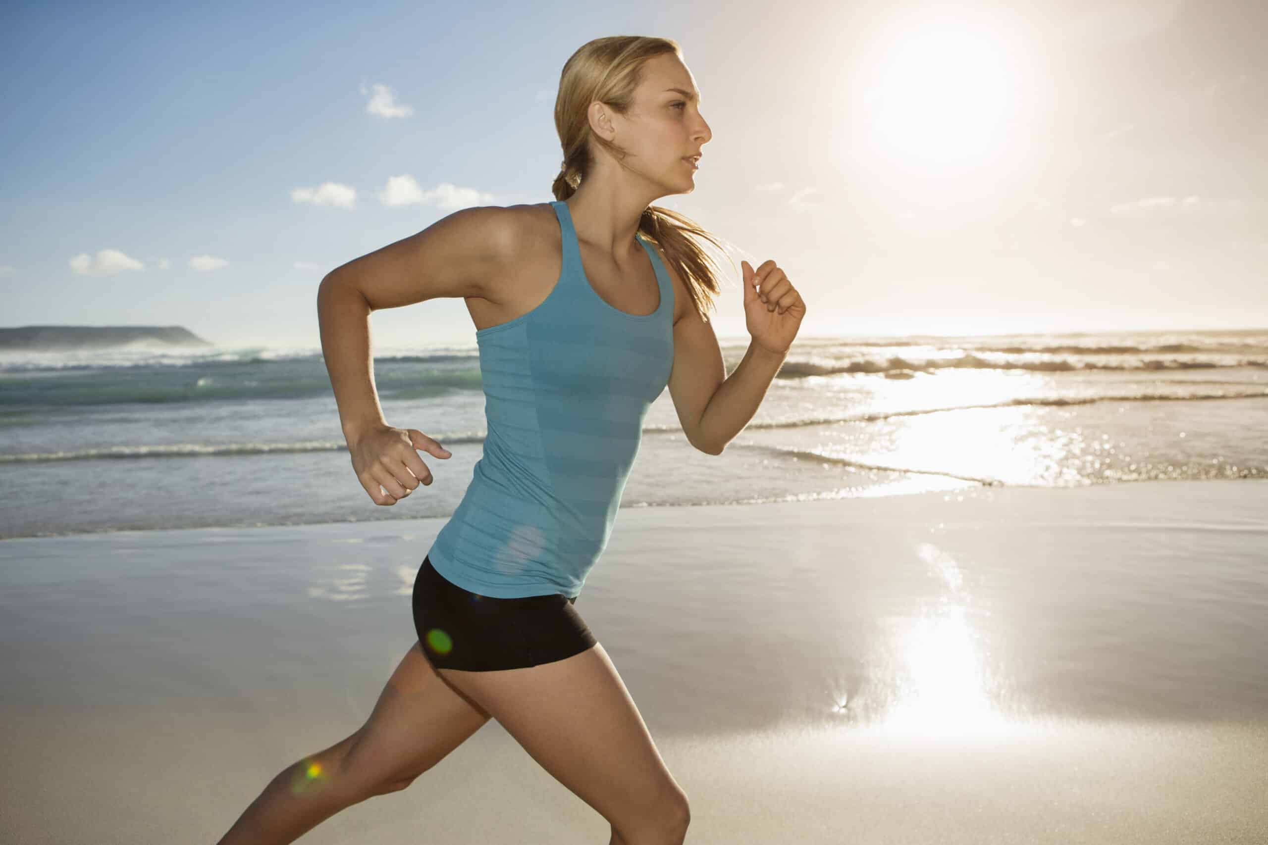 Eine Frau, die bei Sonnenaufgang an einem Strand joggt, bekleidet mit einem blauen Tanktop und schwarzen Shorts, während die Sonne einen warmen Schein auf den nassen Sand wirft und sich darin spiegelt. © Fotografie Tomas Rodriguez