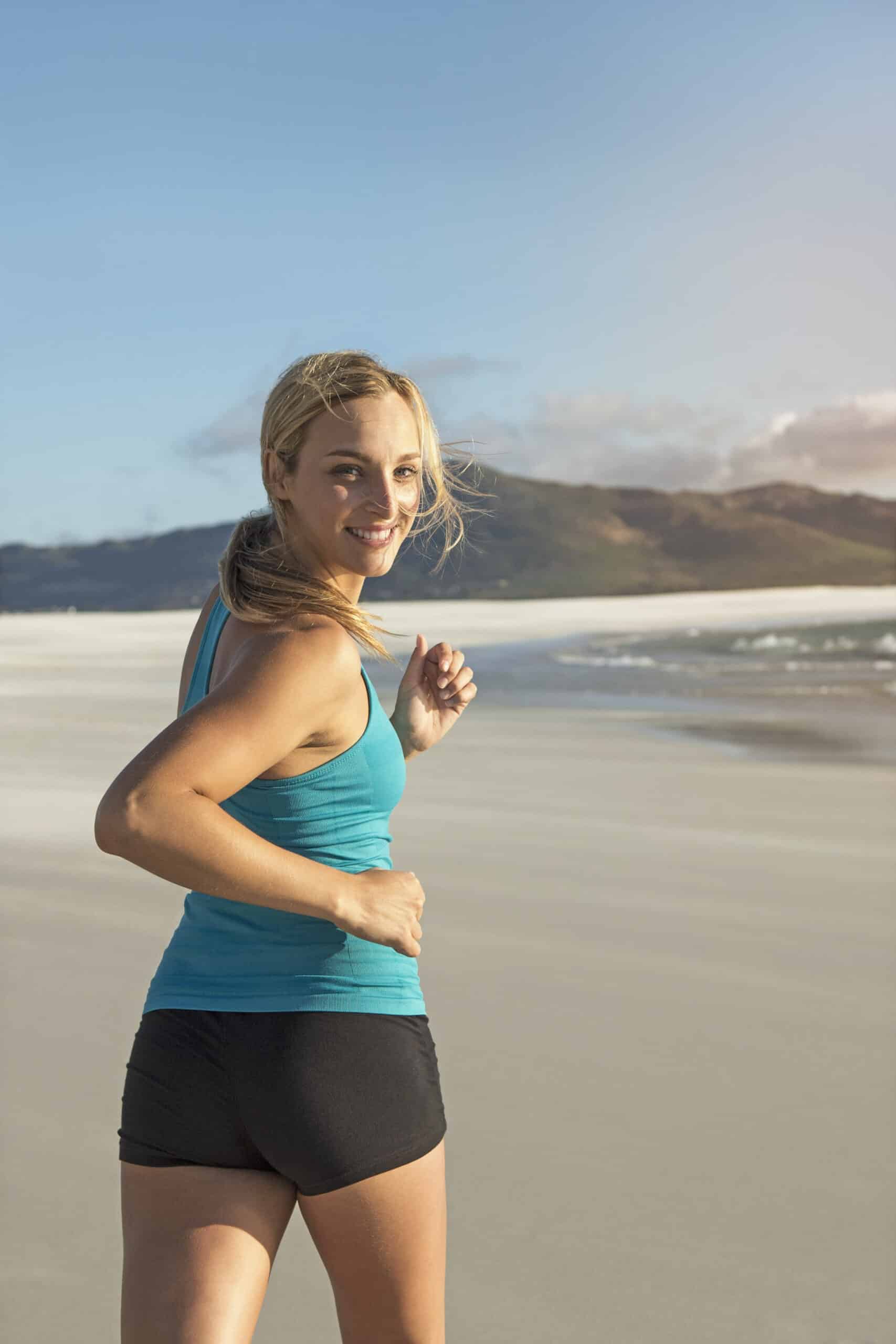 Eine Frau mit blonden Haaren, die ein blaues Tanktop und schwarze Shorts trägt, lächelt beim Joggen an einem Sandstrand mit Bergen im Hintergrund. © Fotografie Tomas Rodriguez