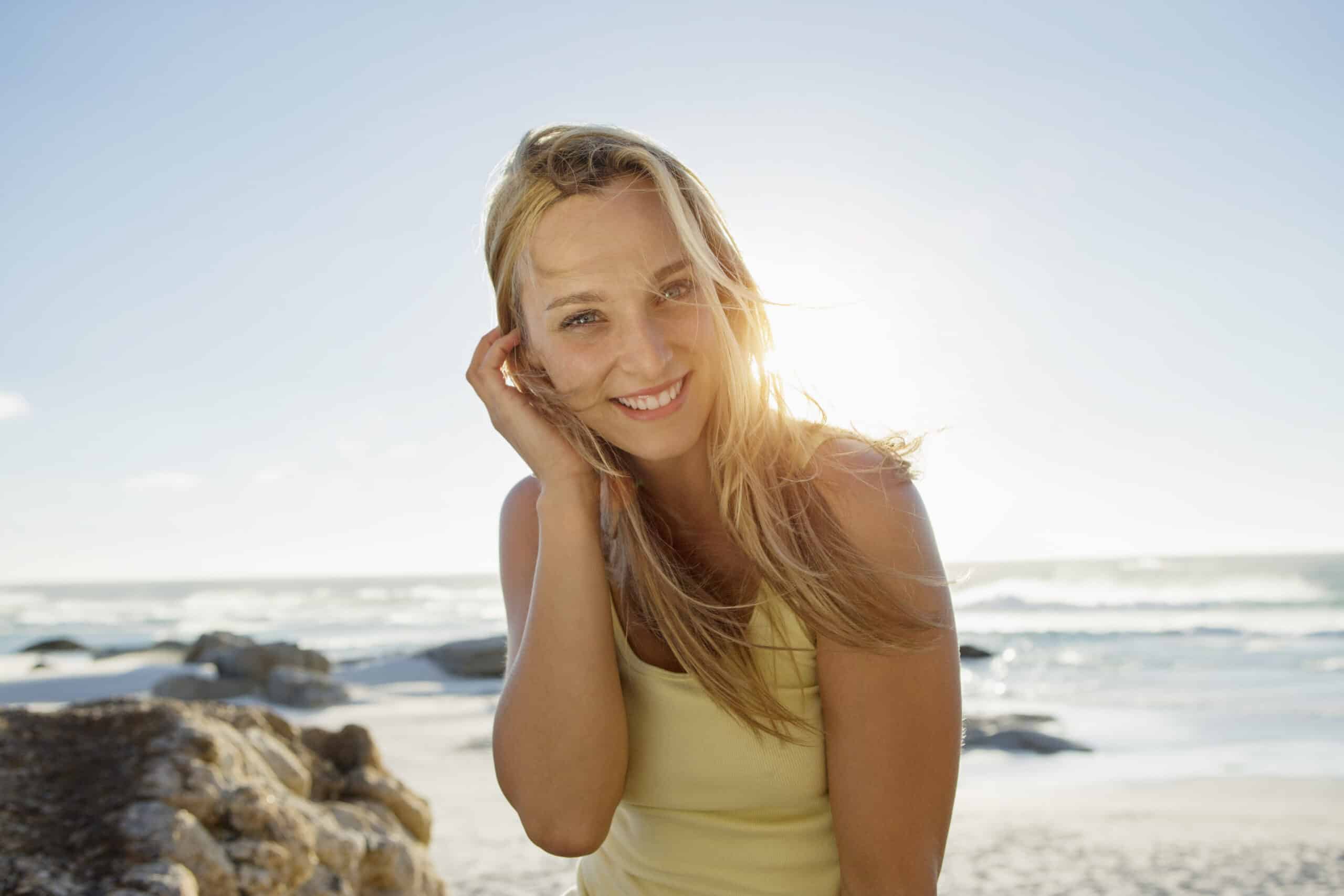 Eine fröhliche Frau lächelt an einem sonnigen Strand in die Kamera, während ihr Haar im Wind weht. Im Hintergrund sind das Meer und die Felsen zu sehen. Sie trägt ein gelbes Oberteil. © Fotografie Tomas Rodriguez