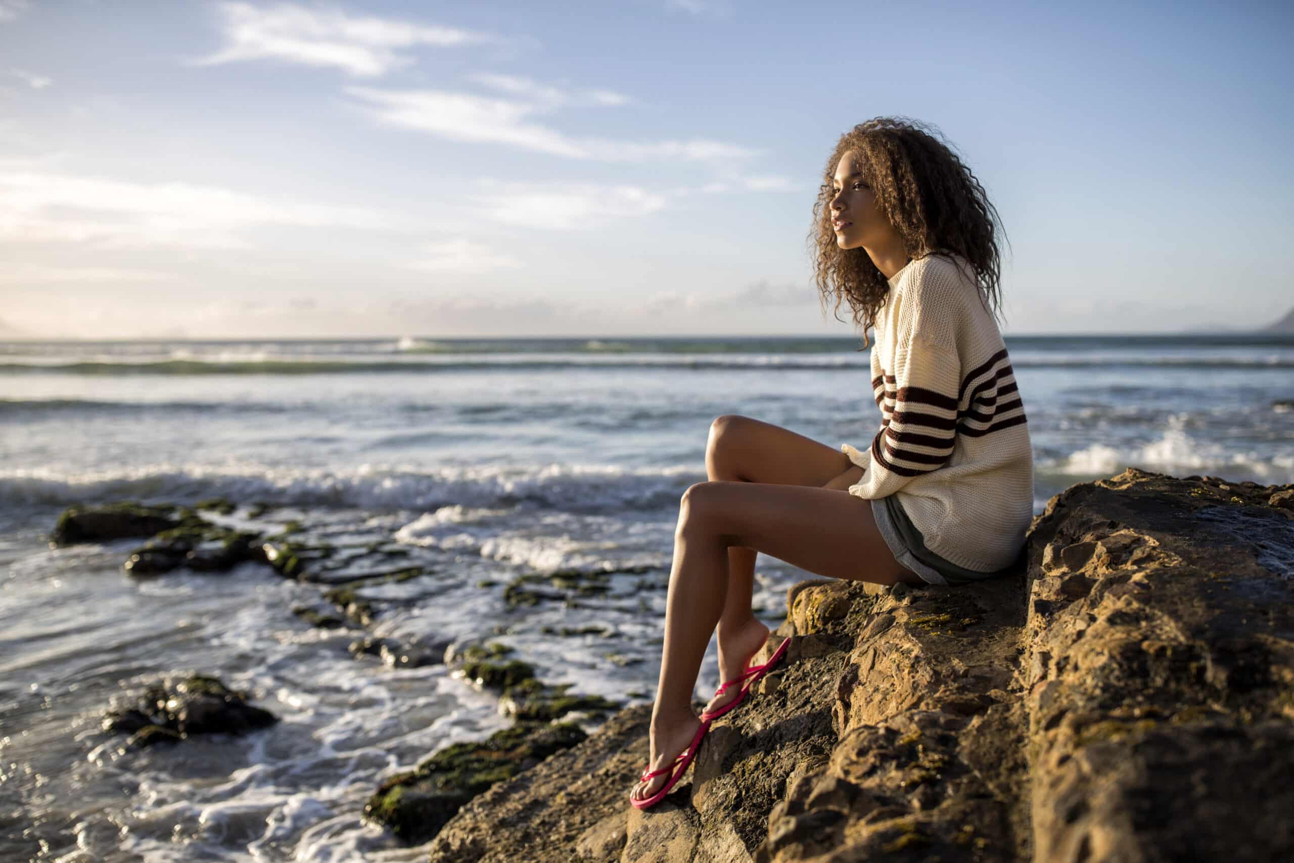Eine Frau sitzt auf einem Felsvorsprung am Meer und blickt auf den Horizont. Sie trägt einen weiß-braun gestreiften Pullover und Shorts sowie rote Sandalen. Im Hintergrund geht die Sonne unter. © Fotografie Tomas Rodriguez