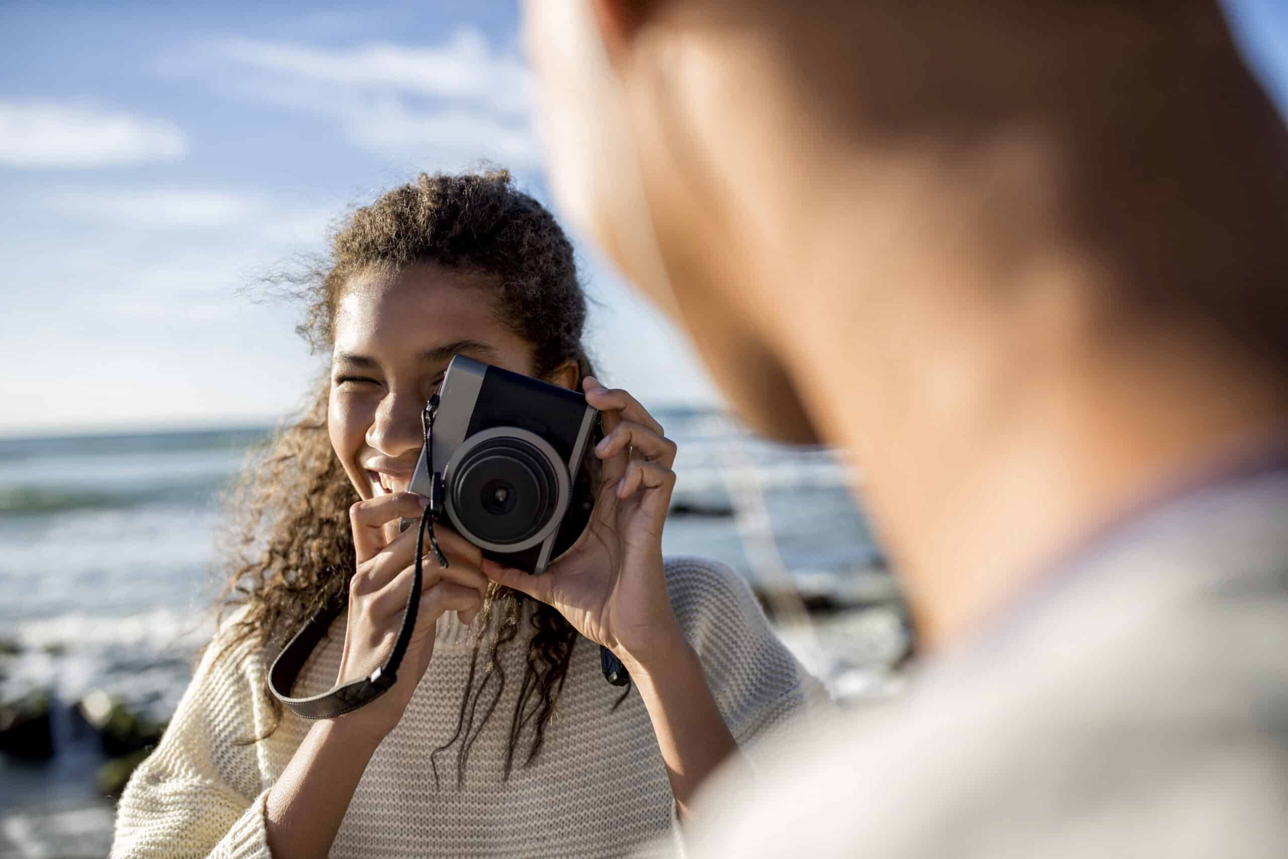 Eine junge Frau mit lockigem Haar fotografiert an einem sonnigen Tag mit einer Digitalkamera eine andere Person im Freien am Meer. © Fotografie Tomas Rodriguez