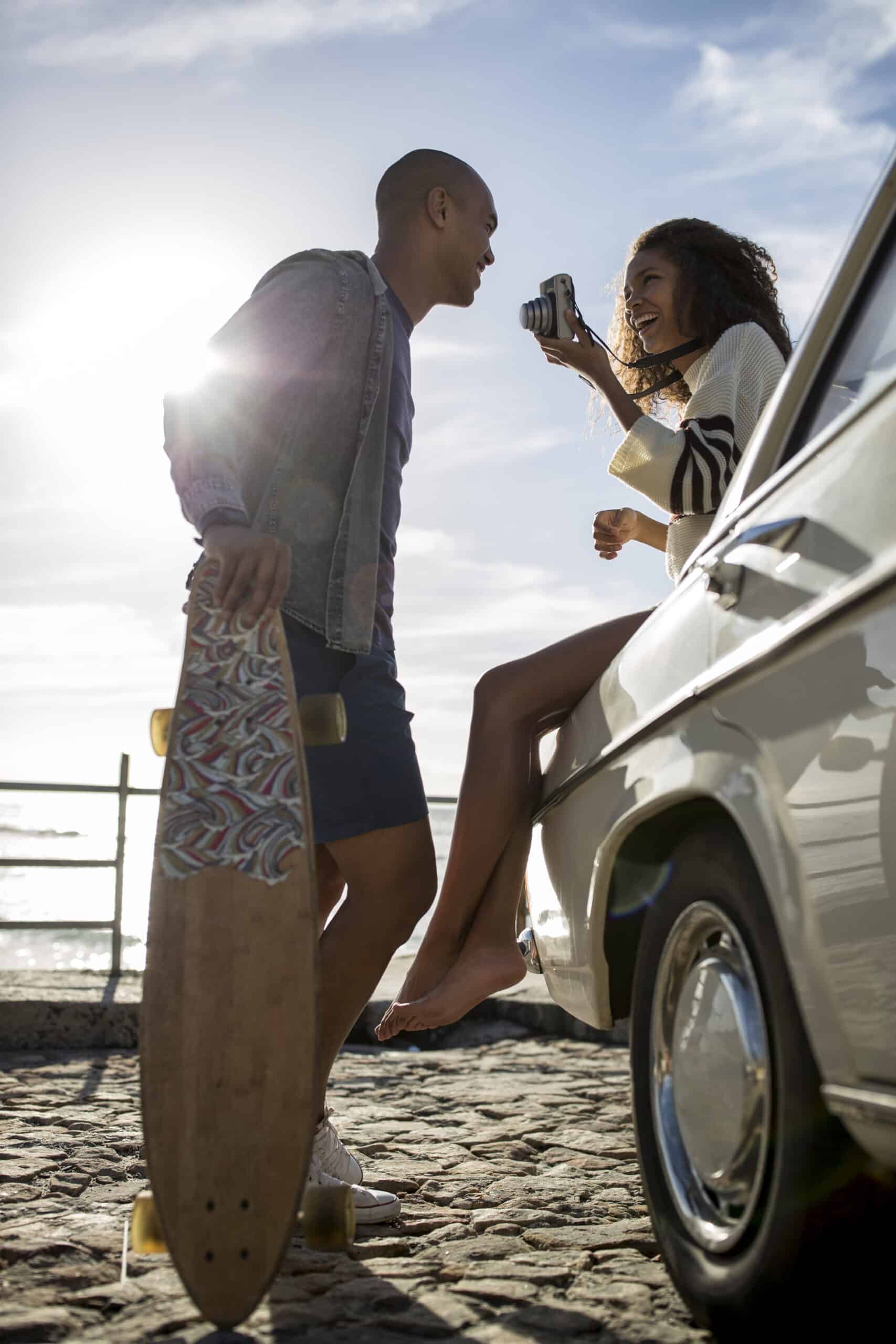 Ein fröhliches Paar genießt einen sonnigen Tag am Meer; die Frau sitzt mit einer Kamera in der Hand auf der Motorhaube eines Autos, während der Mann mit einem Skateboard daneben steht und sich fröhlich unterhält. © Fotografie Tomas Rodriguez
