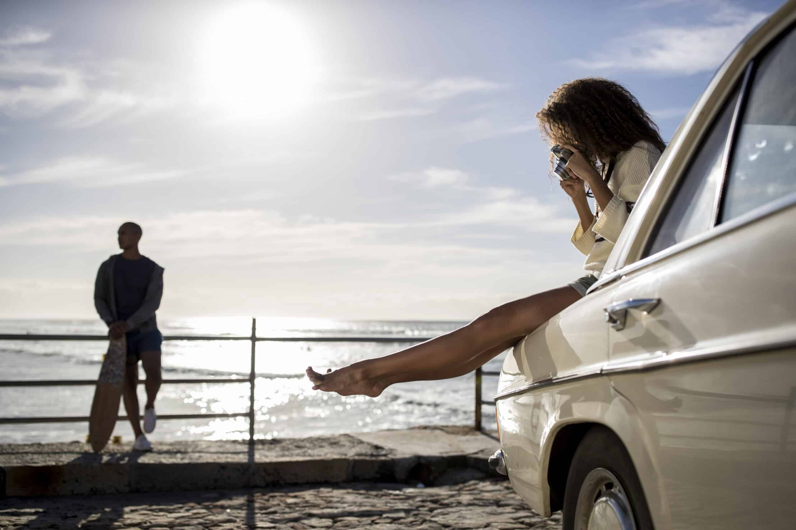 Eine Frau sitzt in einem Oldtimer, streckt ihr Bein aus und macht ein Foto von einem Mann, der unter einem klaren Himmel an der Strandpromenade steht. © Fotografie Tomas Rodriguez