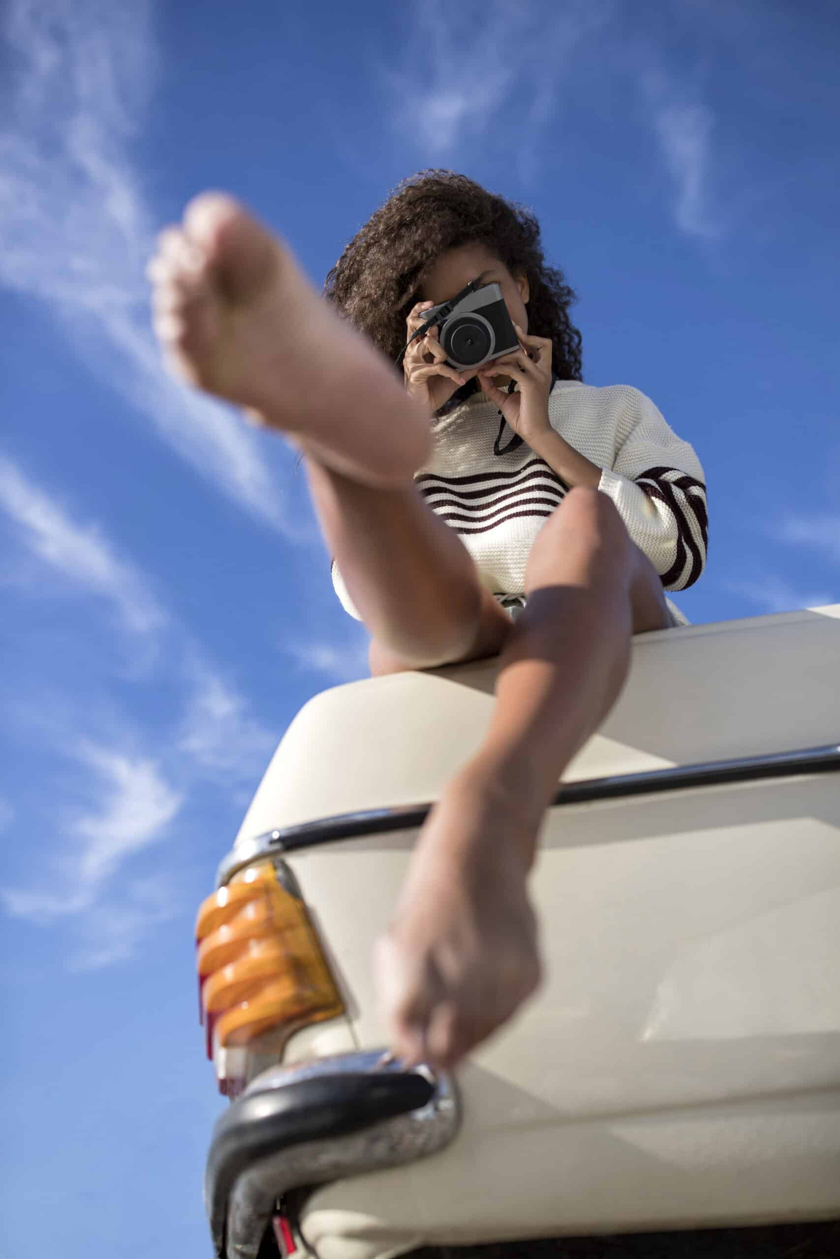 Ein Kind mit lockigem Haar und gestreiftem Hemd sitzt auf dem Dach eines Lieferwagens und macht unter einem klaren blauen Himmel ein Foto mit einer Kamera, den Fuß in Richtung Kamera ausgestreckt. © Fotografie Tomas Rodriguez