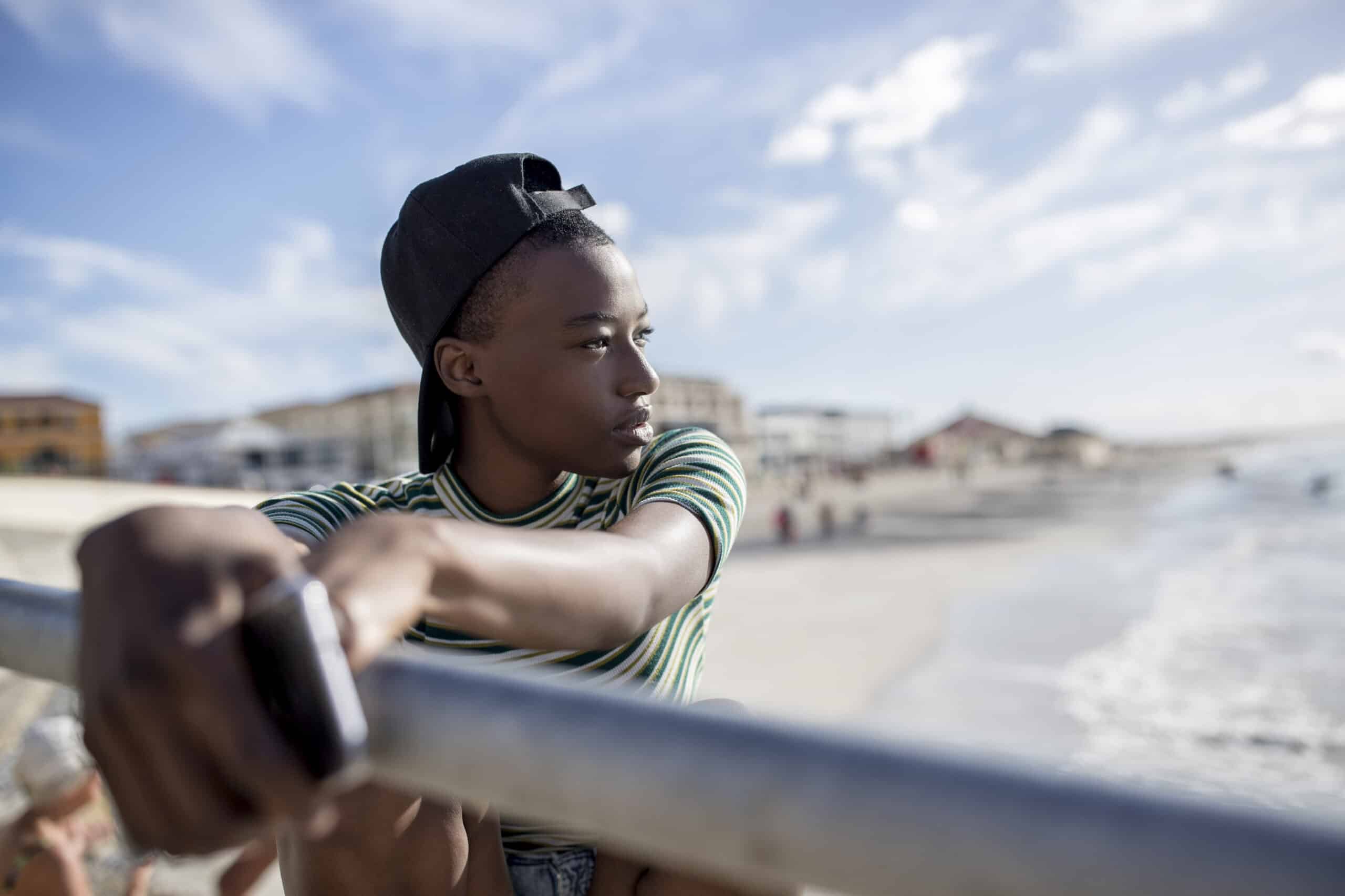 Ein junger Mensch mit verkehrt herum aufgesetzter Kappe lehnt an einem Geländer am Strand und blickt nachdenklich in die Ferne. Im Hintergrund sind Strandbesucher und Gebäude unscharf zu sehen. © Fotografie Tomas Rodriguez