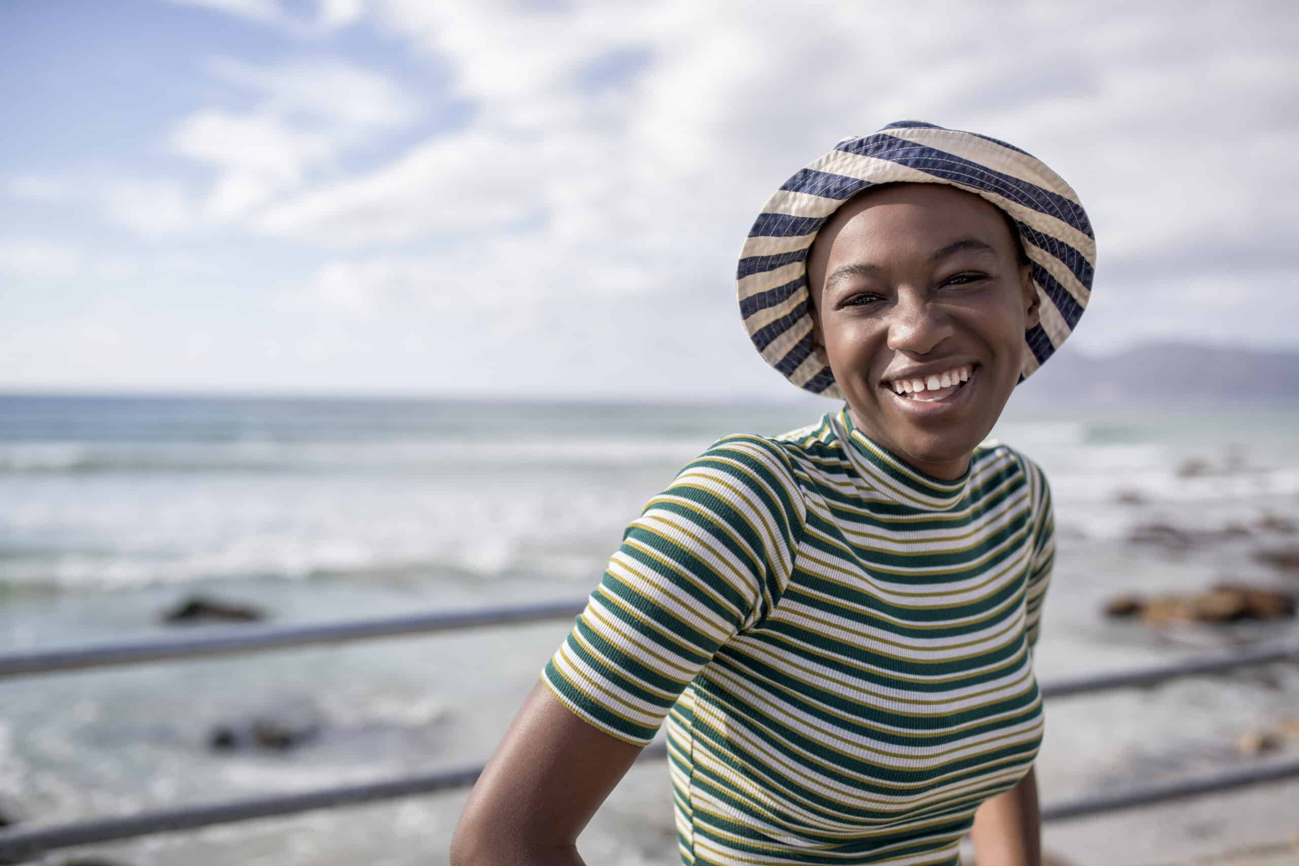 Eine fröhliche junge Frau mit gestreifter Mütze und grün gestreiftem Oberteil lächelt im Freien, mit dem Meer und einem klaren Himmel im Hintergrund. © Fotografie Tomas Rodriguez