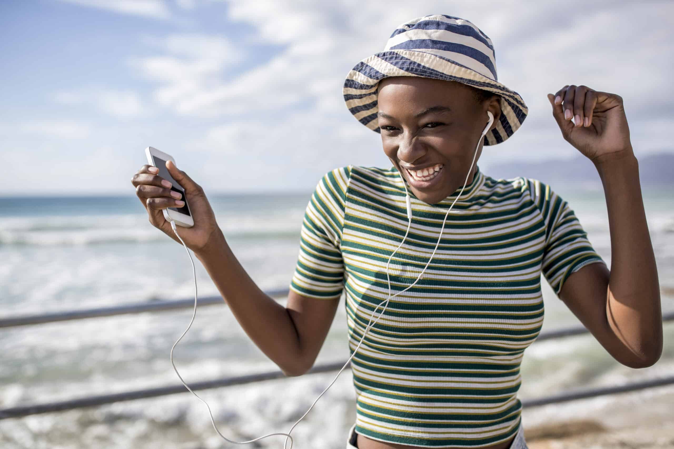 Eine fröhliche junge Frau mit gestreifter Mütze und T-Shirt hört über Kopfhörer Musik auf ihrem Handy und tanzt an einem Geländer am Meer mit dem Meer im Hintergrund. © Fotografie Tomas Rodriguez