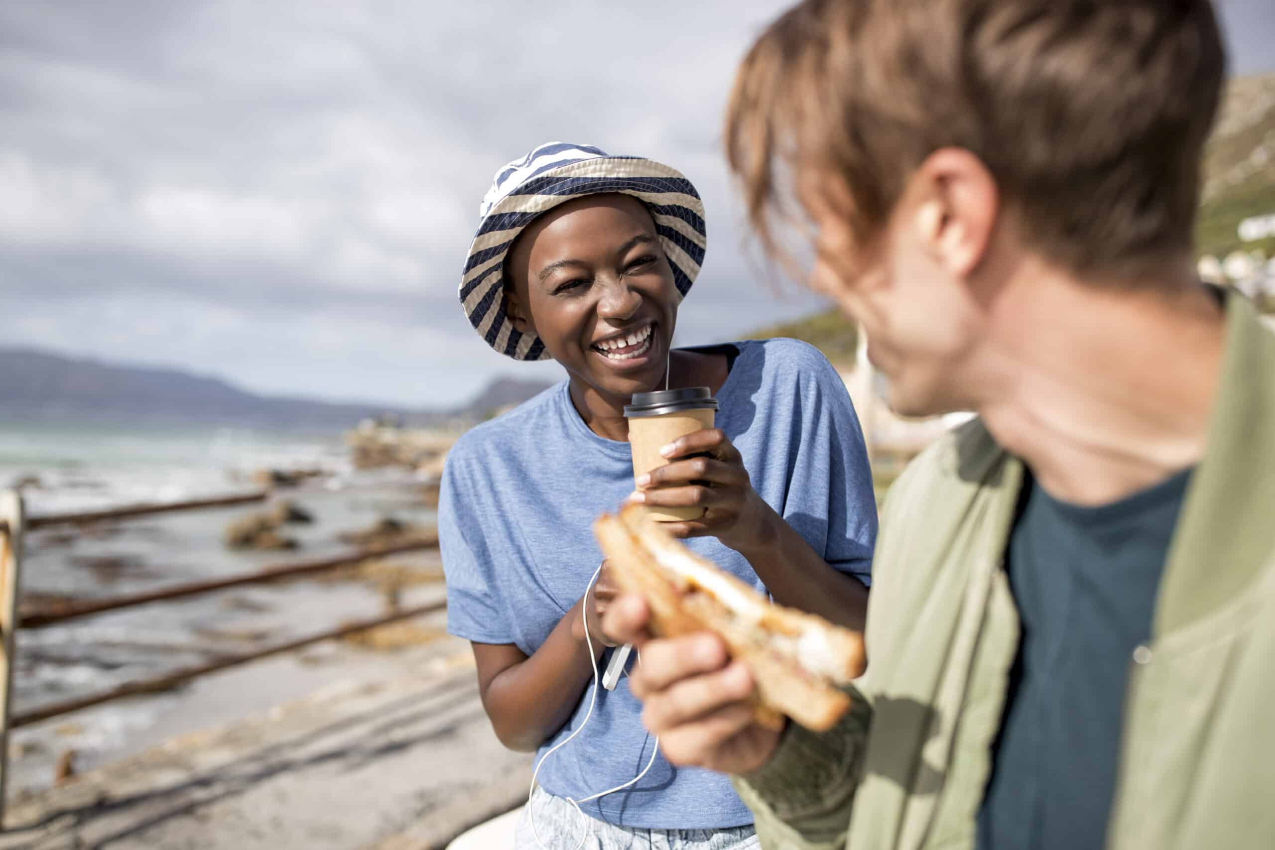 Eine fröhliche Frau mit Hut und einer Kaffeetasse in der Hand unterhält sich mit einem Mann, der an einem sonnigen Strand ein Sandwich isst. © Fotografie Tomas Rodriguez