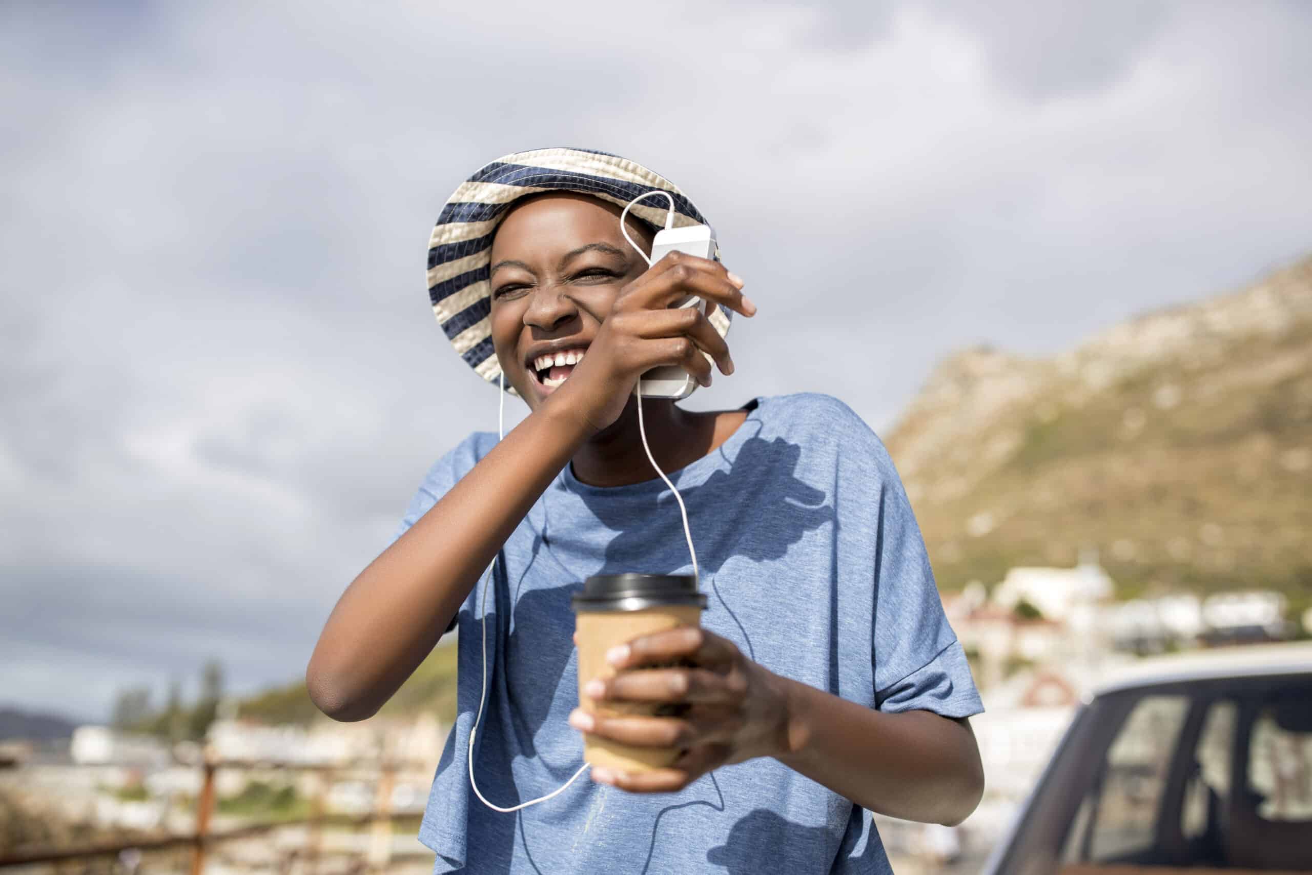Eine fröhliche junge Frau mit gestreifter Mütze und Kopfhörern lacht, während sie im Freien eine Kaffeetasse hält, vor der malerischen Bergkulisse unter einem bewölkten Himmel. © Fotografie Tomas Rodriguez