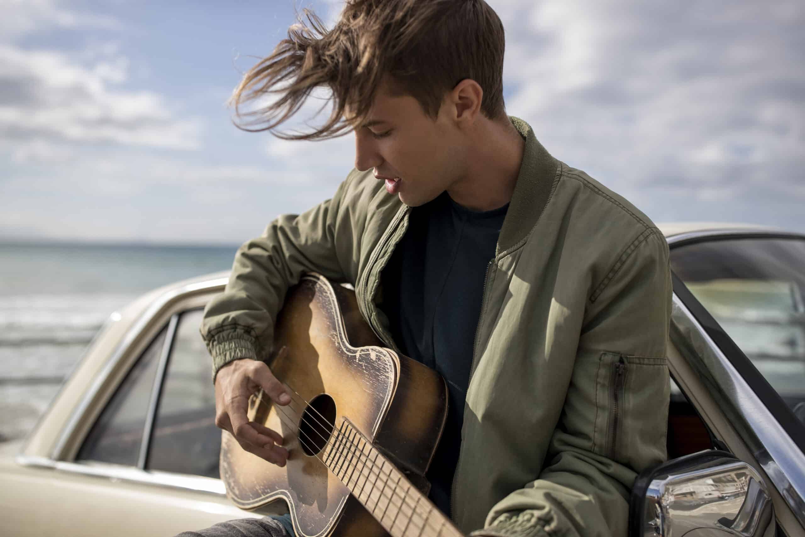 Ein junger Mann spielt Akustikgitarre, sitzt auf der Motorhaube eines Oldtimers am Meer und eine sanfte Brise weht durch seine Haare. © Fotografie Tomas Rodriguez