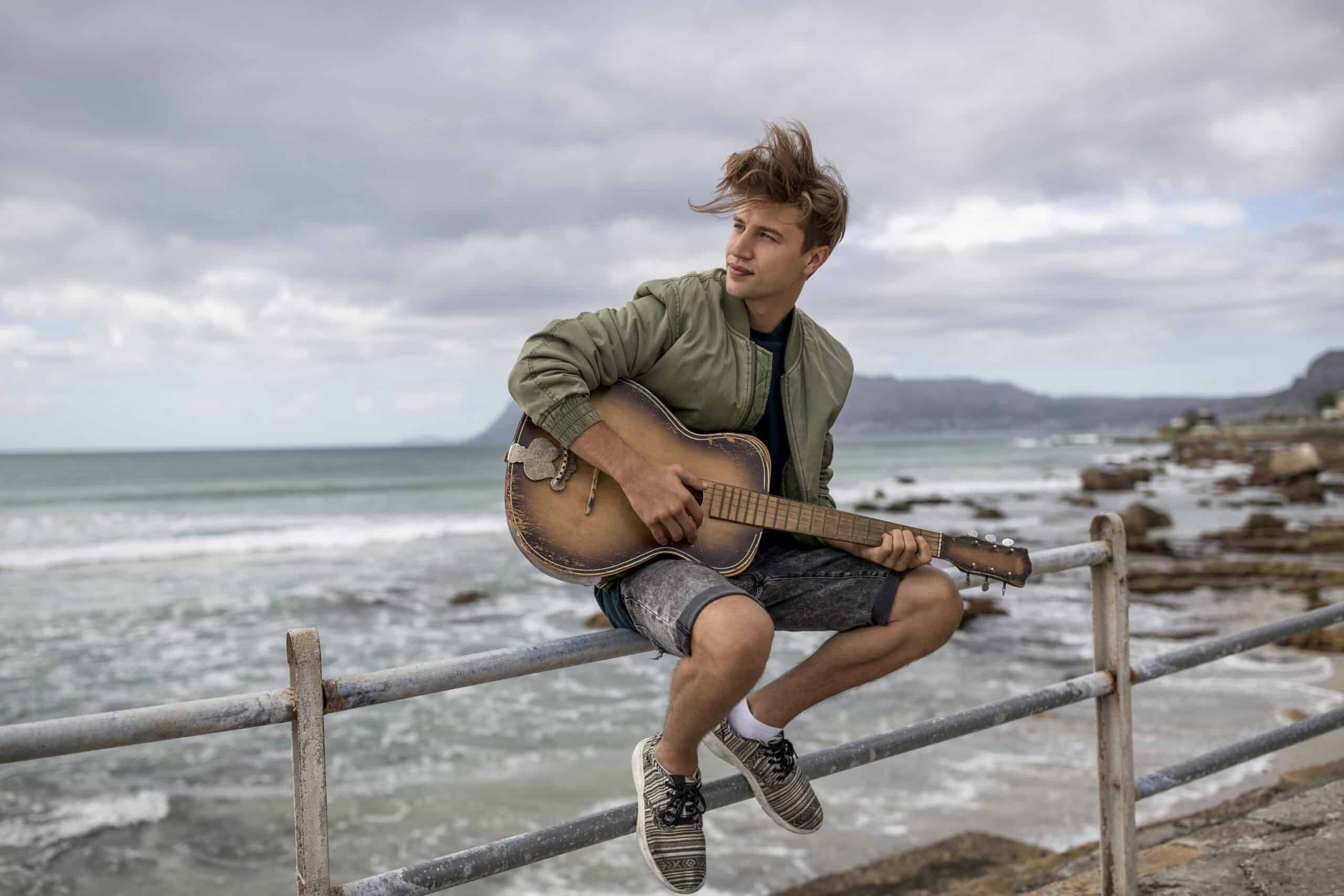 Ein junger Mann sitzt auf einem Geländer am Meer und hält eine Gitarre, im Hintergrund ein dramatischer Wolkenhimmel und eine felsige Küste. © Fotografie Tomas Rodriguez