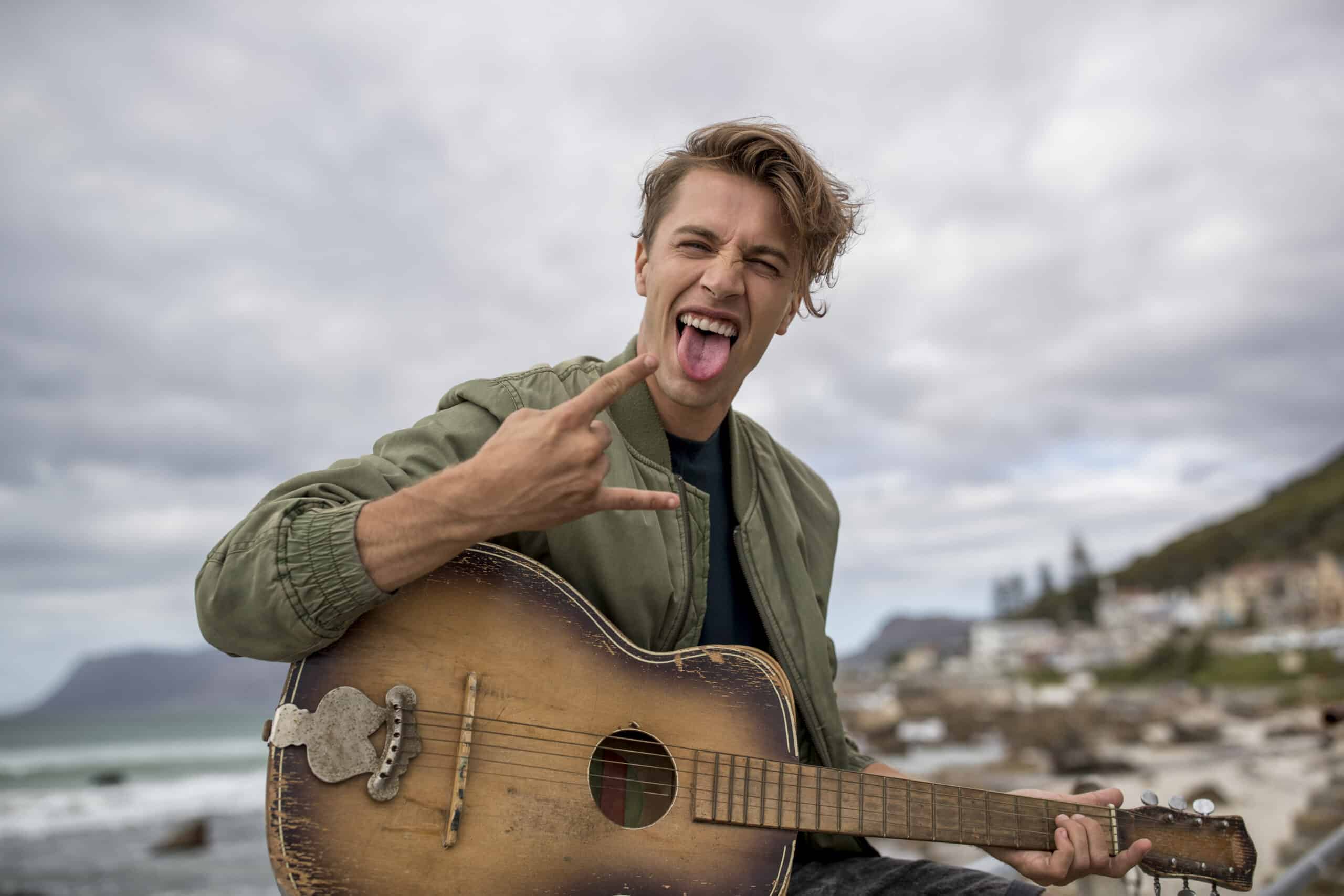 Ein fröhlicher junger Mann mit zerzaustem Haar spielt an einem Strand eine Akustikgitarre, macht eine verspielte Rock-and-Roll-Handbewegung und streckt die Zunge heraus, im Hintergrund sind bewölkter Himmel und eine Küstenlinie zu sehen. © Fotografie Tomas Rodriguez