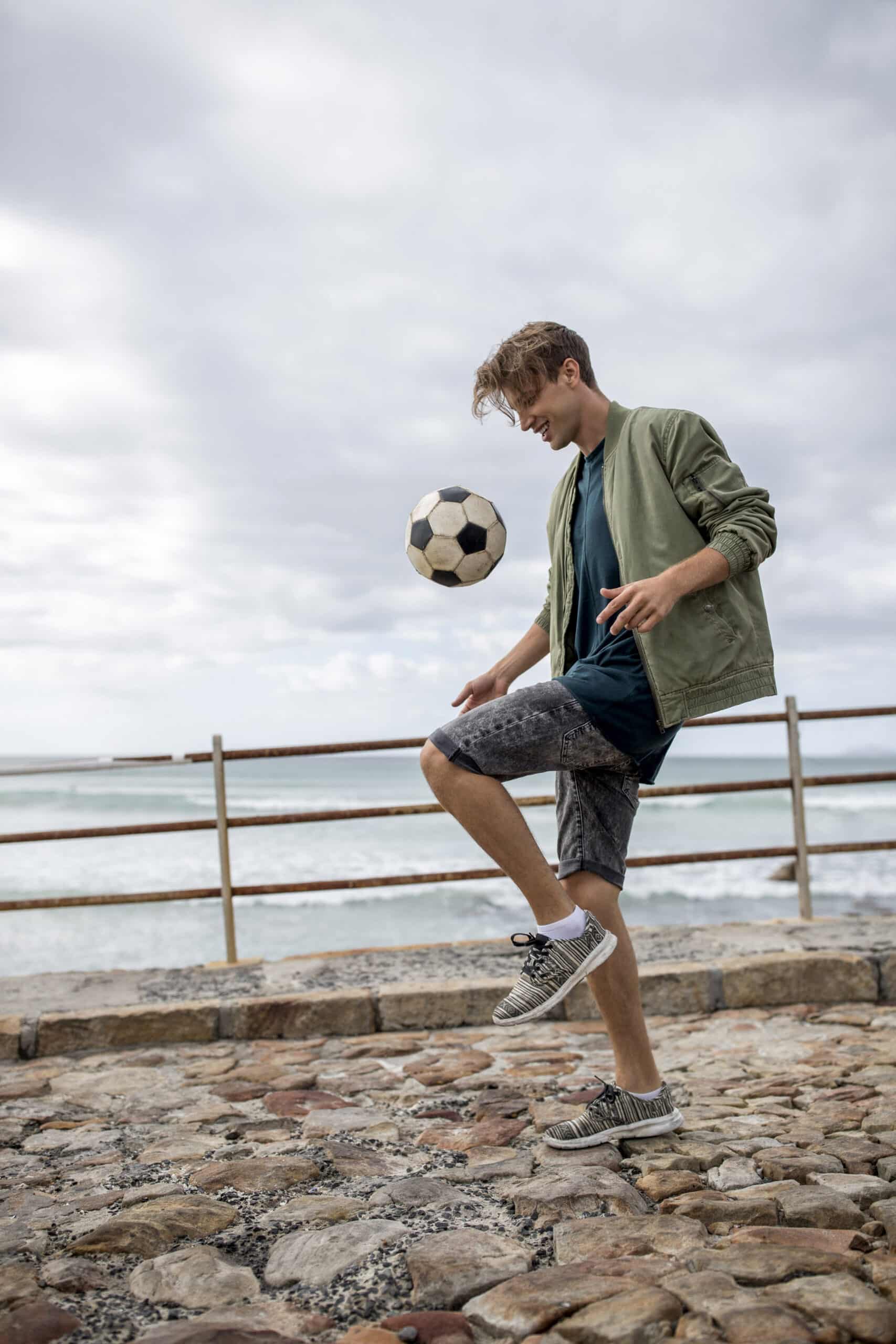 Ein junger Mann jongliert mit einem Fußball auf einem steinigen Strandweg in der Nähe eines Geländers, im Hintergrund das Meer und ein bewölkter Himmel. © Fotografie Tomas Rodriguez