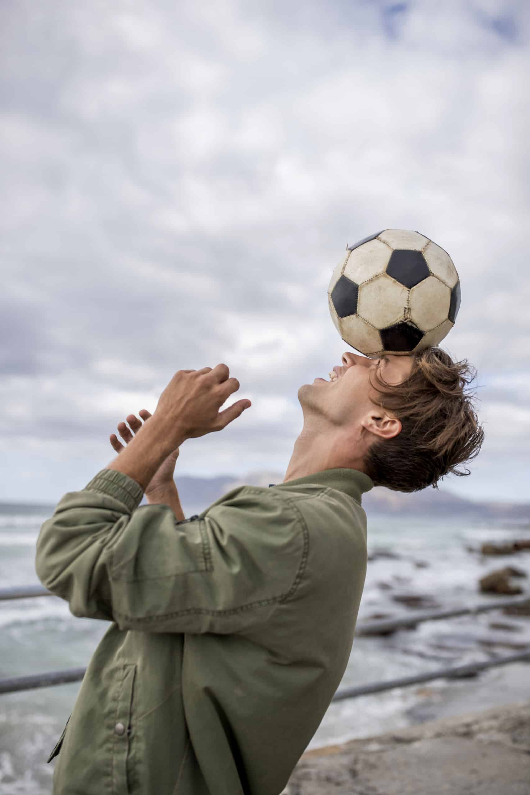 Ein junger Mann balanciert einen Fußball auf seiner Stirn am Meer, über ihm ist ein bewölkter Himmel und im Hintergrund sind Felsen zu sehen. Er trägt eine grüne Jacke und blickt aufmerksam nach oben. © Fotografie Tomas Rodriguez