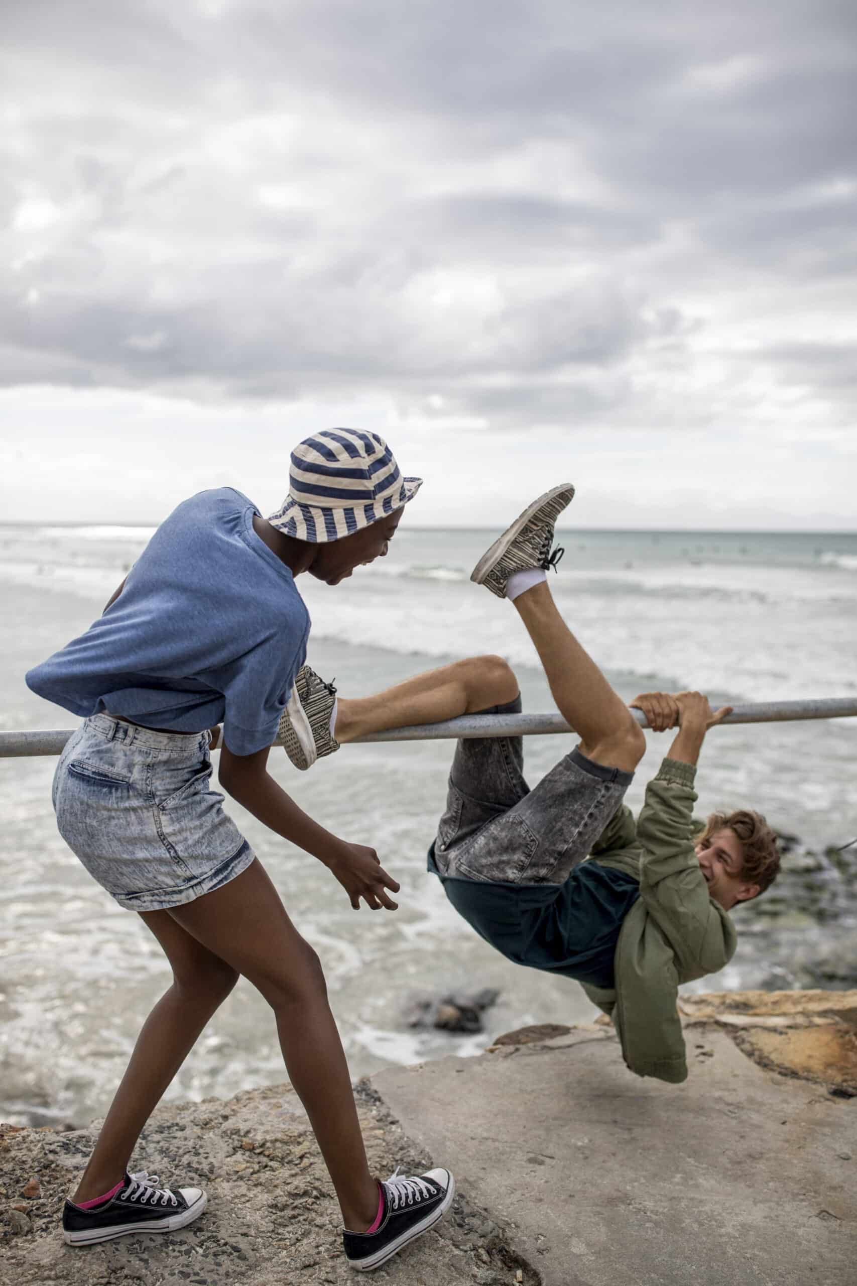 Zwei junge Leute haben an einem bewölkten Tag am Strand Spaß; einer hängt kopfüber an einer Stange, während der andere steht und so tut, als würde er sie spielerisch treten. © Fotografie Tomas Rodriguez