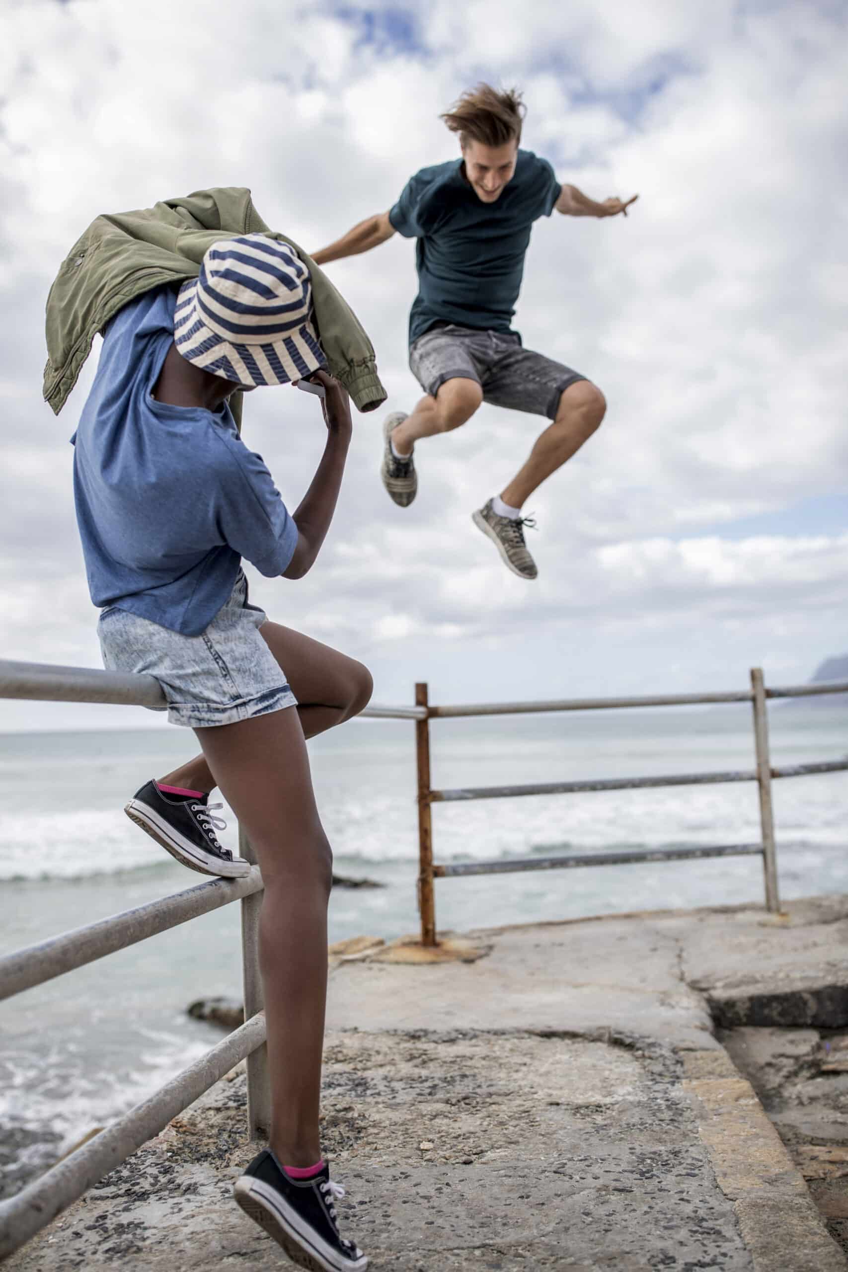 Ein kleiner Junge springt über ein Metallgeländer am Meer, während sich ein anderer Junge mit seinem Hut in der Hand zum Sprung bereit macht. Im Hintergrund brechen Meereswellen unter einem bewölkten Himmel. © Fotografie Tomas Rodriguez