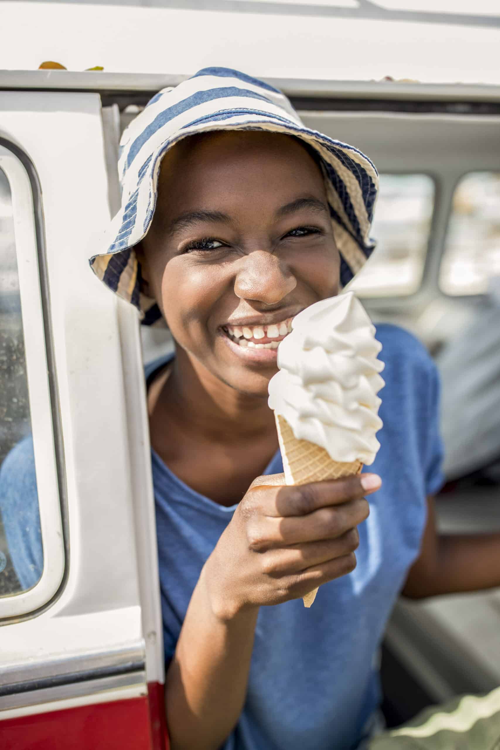 Eine fröhliche junge Frau mit gestreifter Mütze hält eine Vanilleeistüte und lächelt strahlend neben einem Auto an einem sonnigen Tag. © Fotografie Tomas Rodriguez