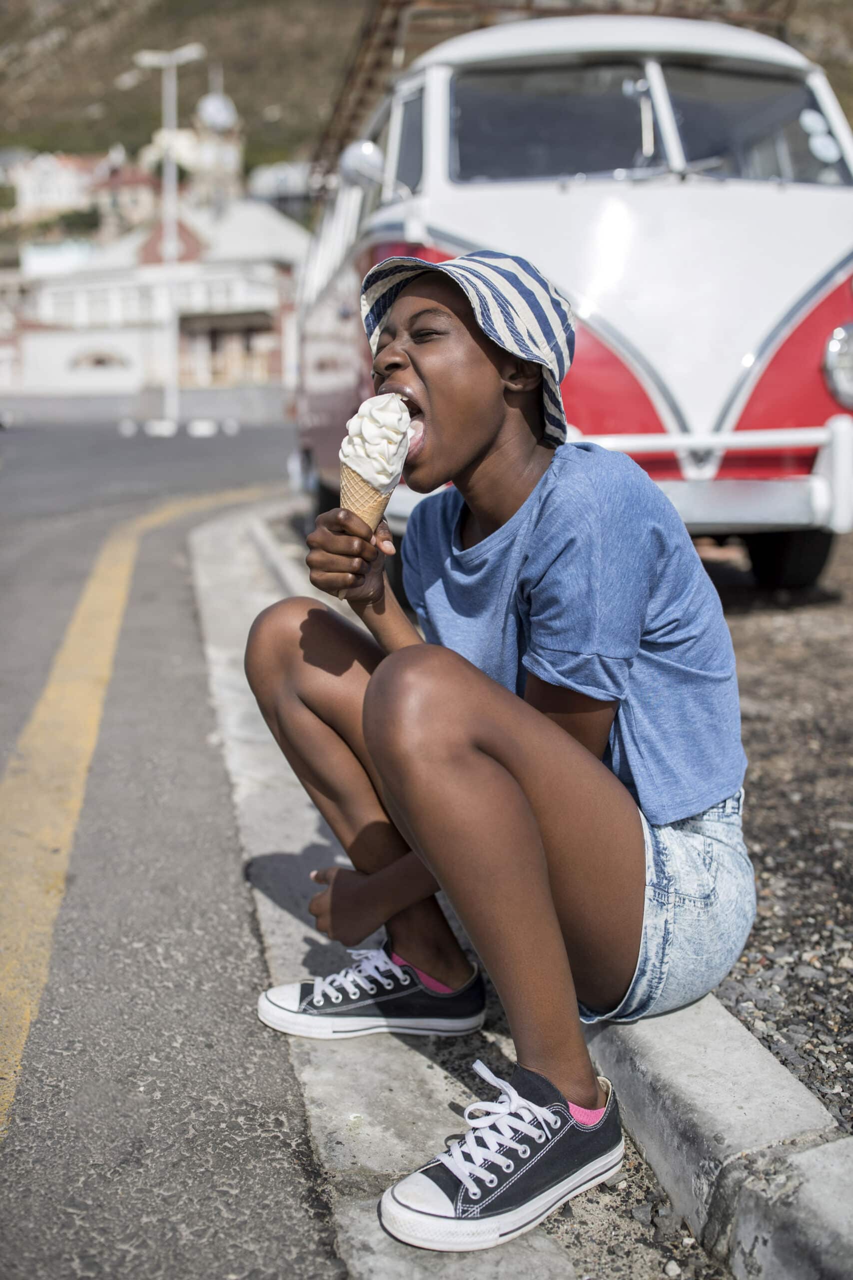 Eine Frau in Freizeitkleidung genießt eine Vanilleeistüte, während sie auf einem Bordstein hockt. An einem sonnigen Tag parkt in der Nähe ein weißer und roter Oldtimer-Van. © Fotografie Tomas Rodriguez