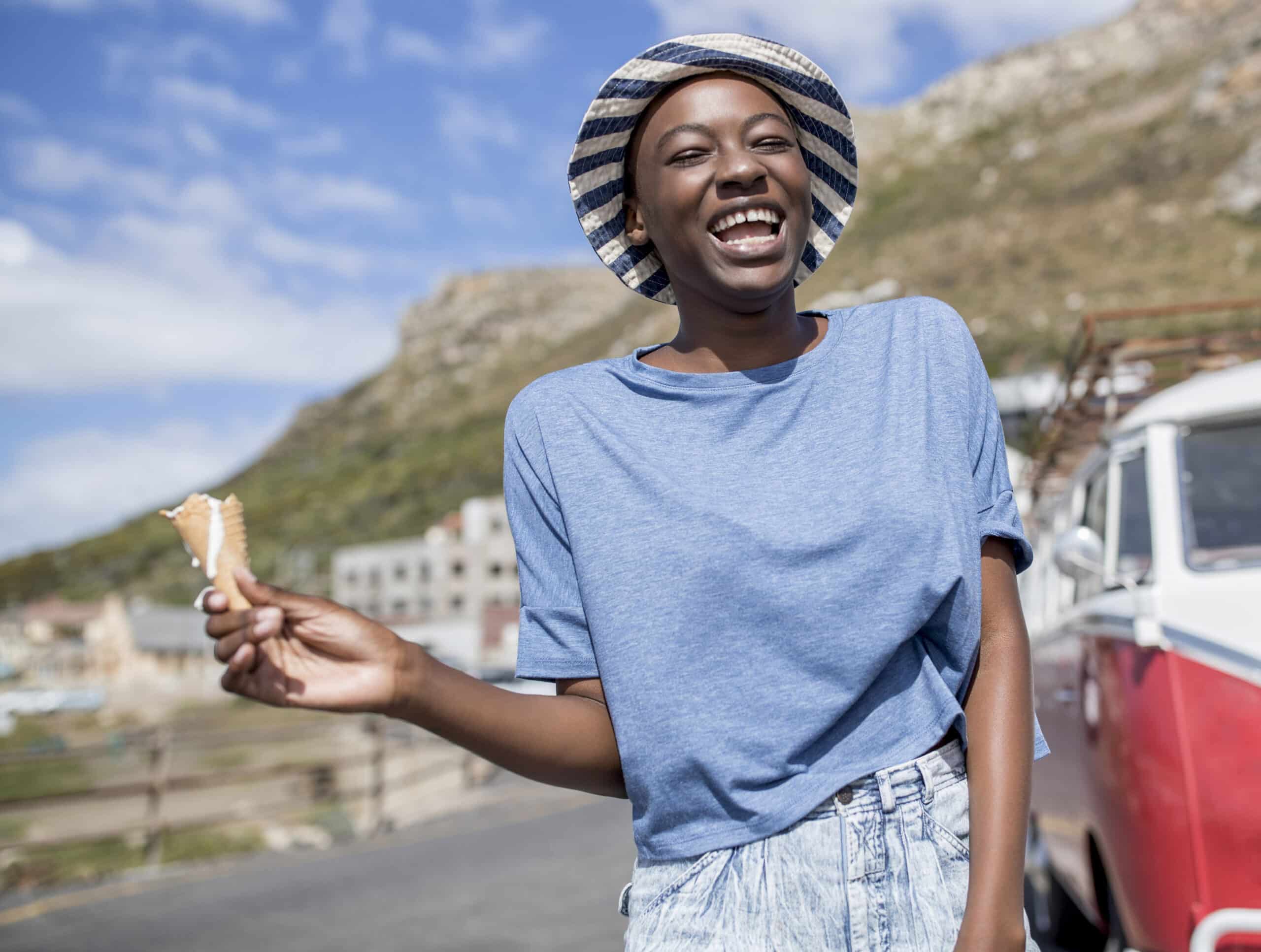 Eine fröhliche Frau mit gestreifter Mütze und blauem T-Shirt hält an einem sonnigen Tag eine Eistüte, im Hintergrund sind Berge und ein Oldtimer-Van zu sehen. © Fotografie Tomas Rodriguez