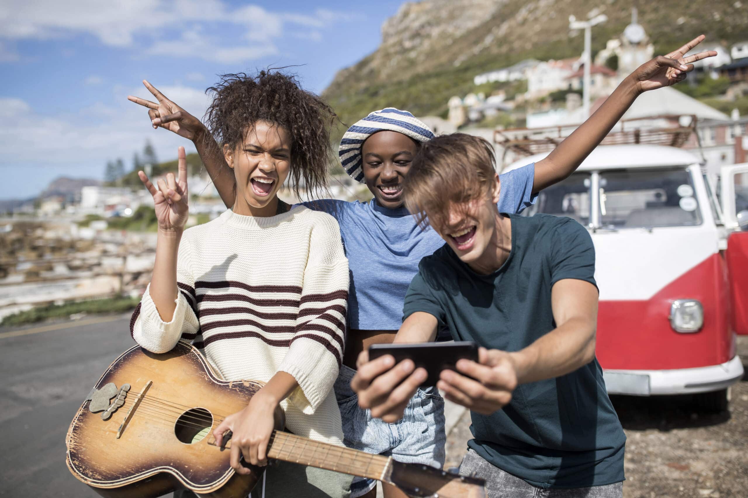 Drei fröhliche Freunde amüsieren sich im Freien in der Nähe eines Lieferwagens. Einer spielt Gitarre, ein anderer macht ein Selfie und der dritte jubelt. Sie befinden sich in der Nähe einer Bergregion unter einem klaren Himmel. © Fotografie Tomas Rodriguez