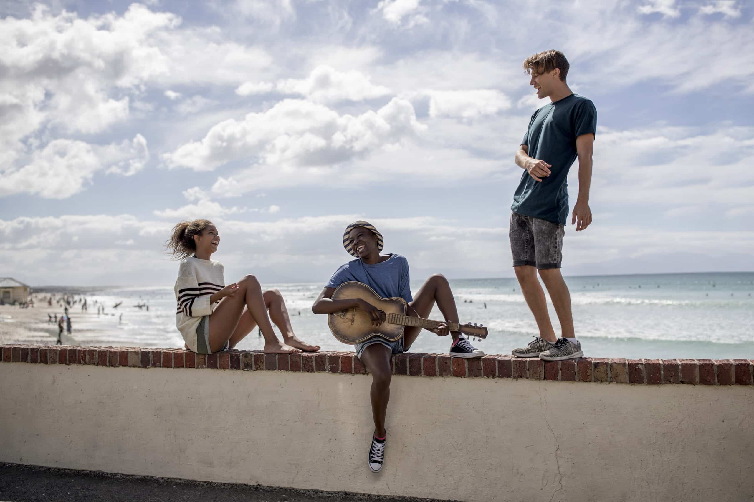 Drei Freunde genießen einen sonnigen Tag am Strand; einer spielt Gitarre, während die anderen zuhören, mit dem Meer und den Strandbesuchern im Hintergrund. © Fotografie Tomas Rodriguez