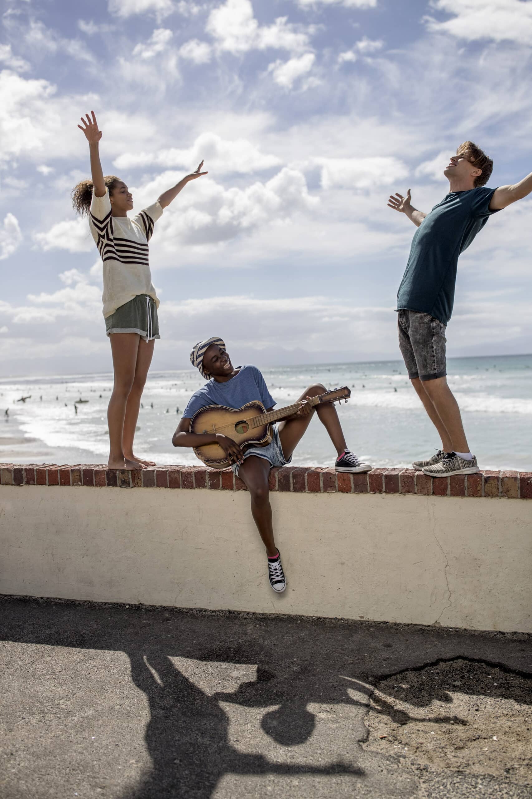 Drei Freunde vergnügen sich am Strand. Zwei stehen auf einer Mauer und strecken sich nach oben, während einer sitzt und Gitarre spielt. Im Hintergrund sehen Sie einen bewölkten Himmel und ein ruhiges Meer. © Fotografie Tomas Rodriguez